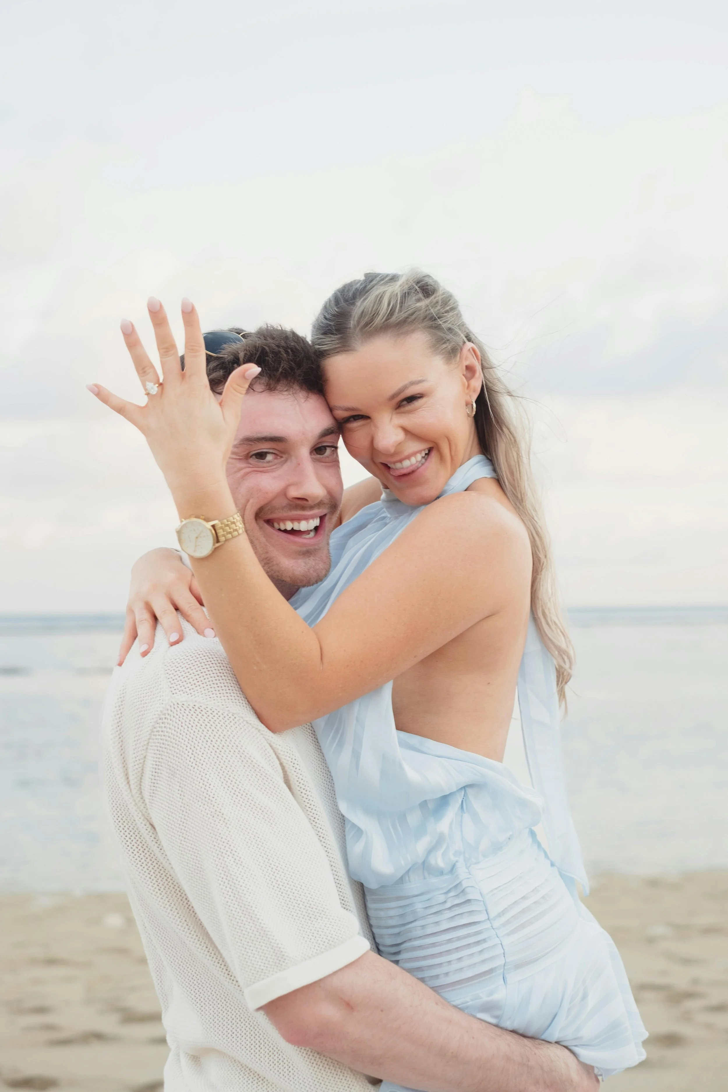 A smiling couple embracing on the beach, the woman showing wedding rings, both wearing light-colored clothing.