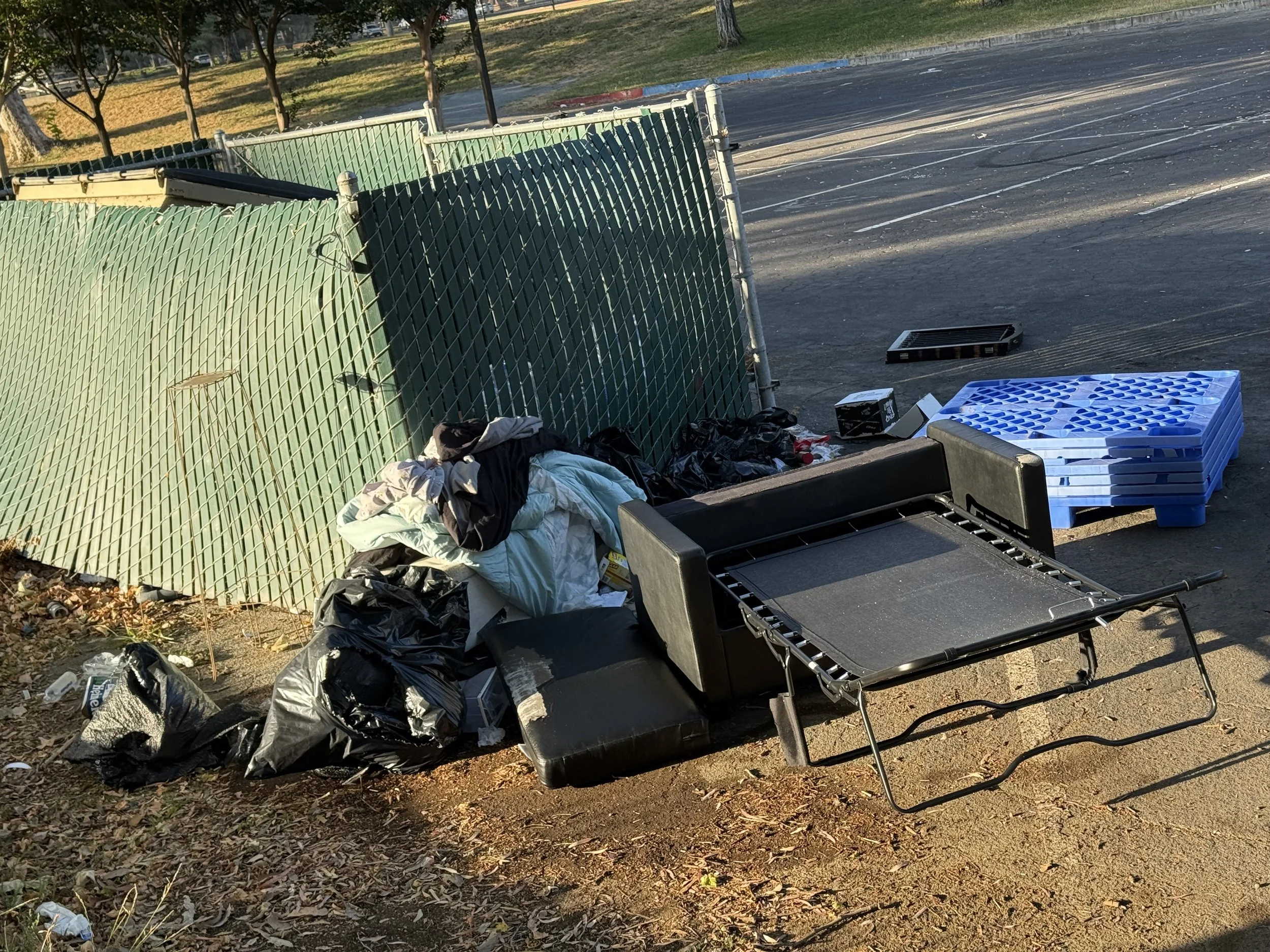 Pile of trash and clutter including clothes, plastic bags, an overturned shopping cart, and a blue plastic pallet next to a green metal fence in a parking lot.