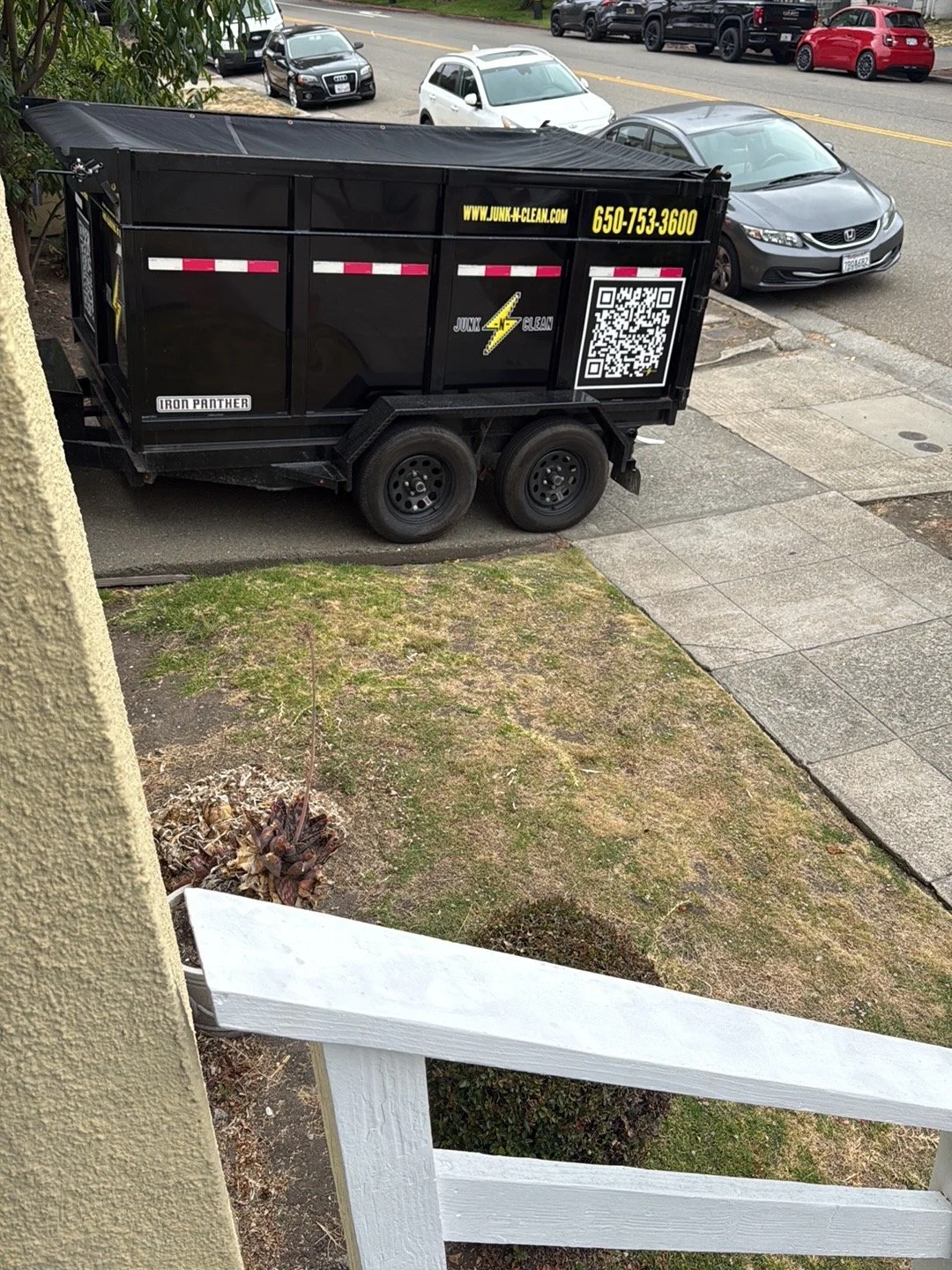 A black junk disposal trailer parked on a sidewalk, with a website and phone number on the side, on a city street with multiple parked cars.