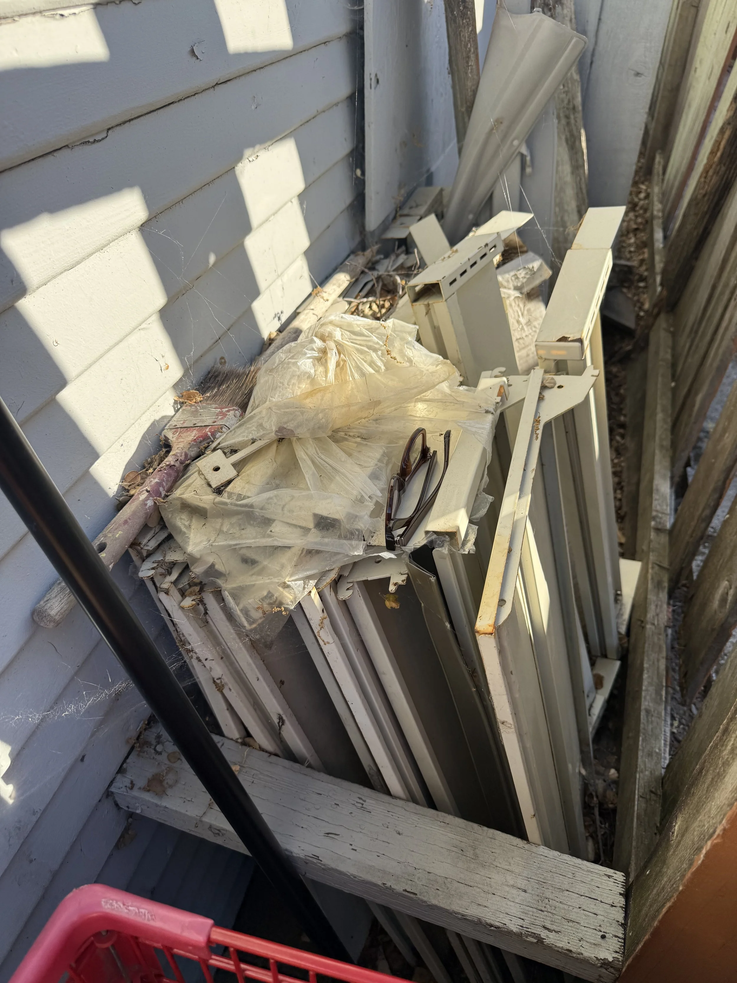 Cluttered outdoor space with discarded metal and plastic objects, including pieces of siding, a plastic bag, glasses, and a broom, next to a light-colored wooden fence and siding.