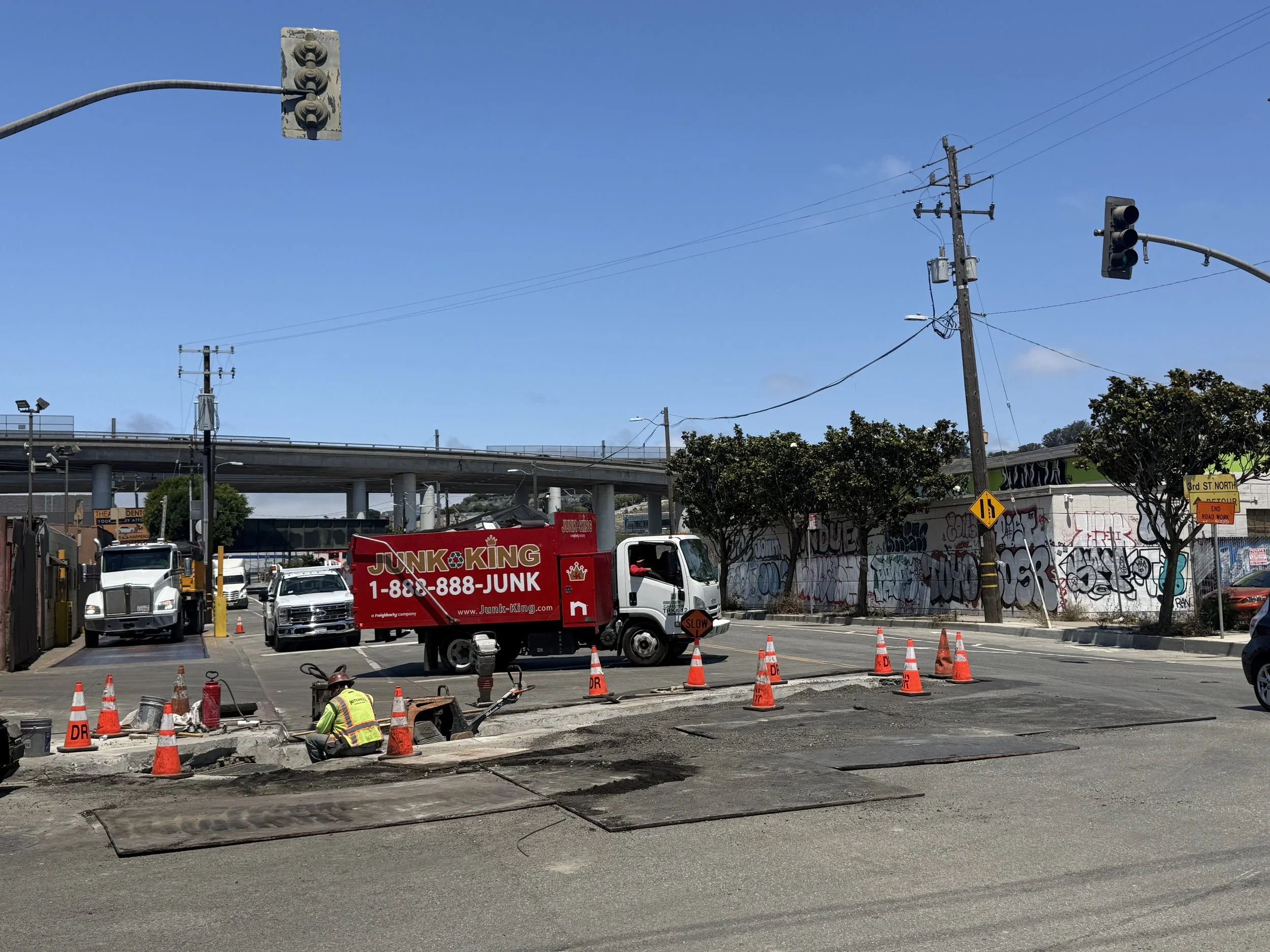 Construction workers working on a street repair or excavation site with orange cones, a JUNK KING truck, and construction equipment, with traffic signals and utility poles in the background.