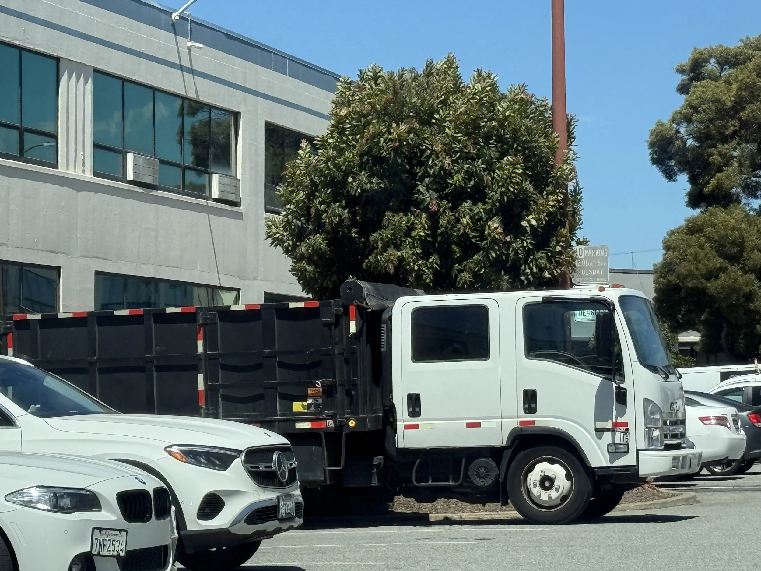 Street scene with parked cars including a white sports car and a truck, trees, and a building with windows under clear blue sky.
