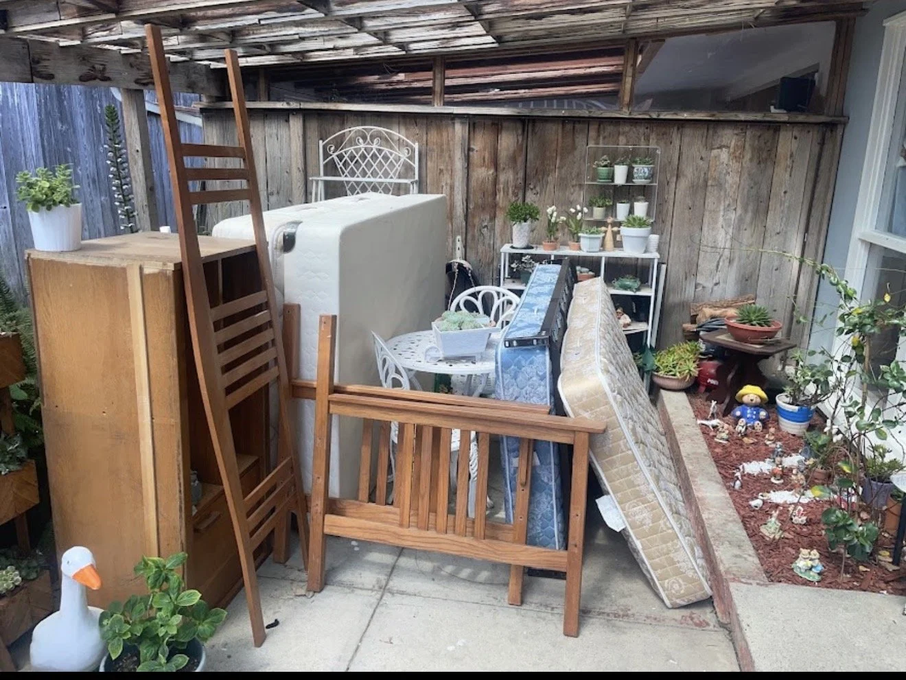 Cluttered backyard storage area with stacked furniture, plants, and miscellaneous items.