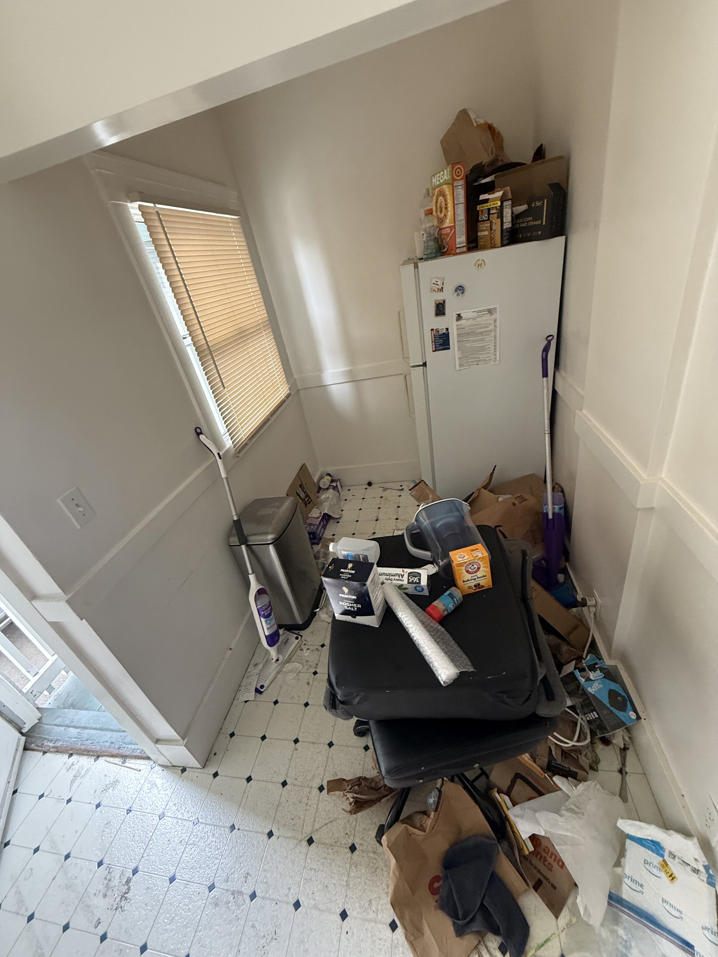 A cluttered room with items on the floor including cardboard boxes, paper bags, and cleaning supplies. There is a white refrigerator against the wall, a window with closed blinds, and a black chair with items on it.
