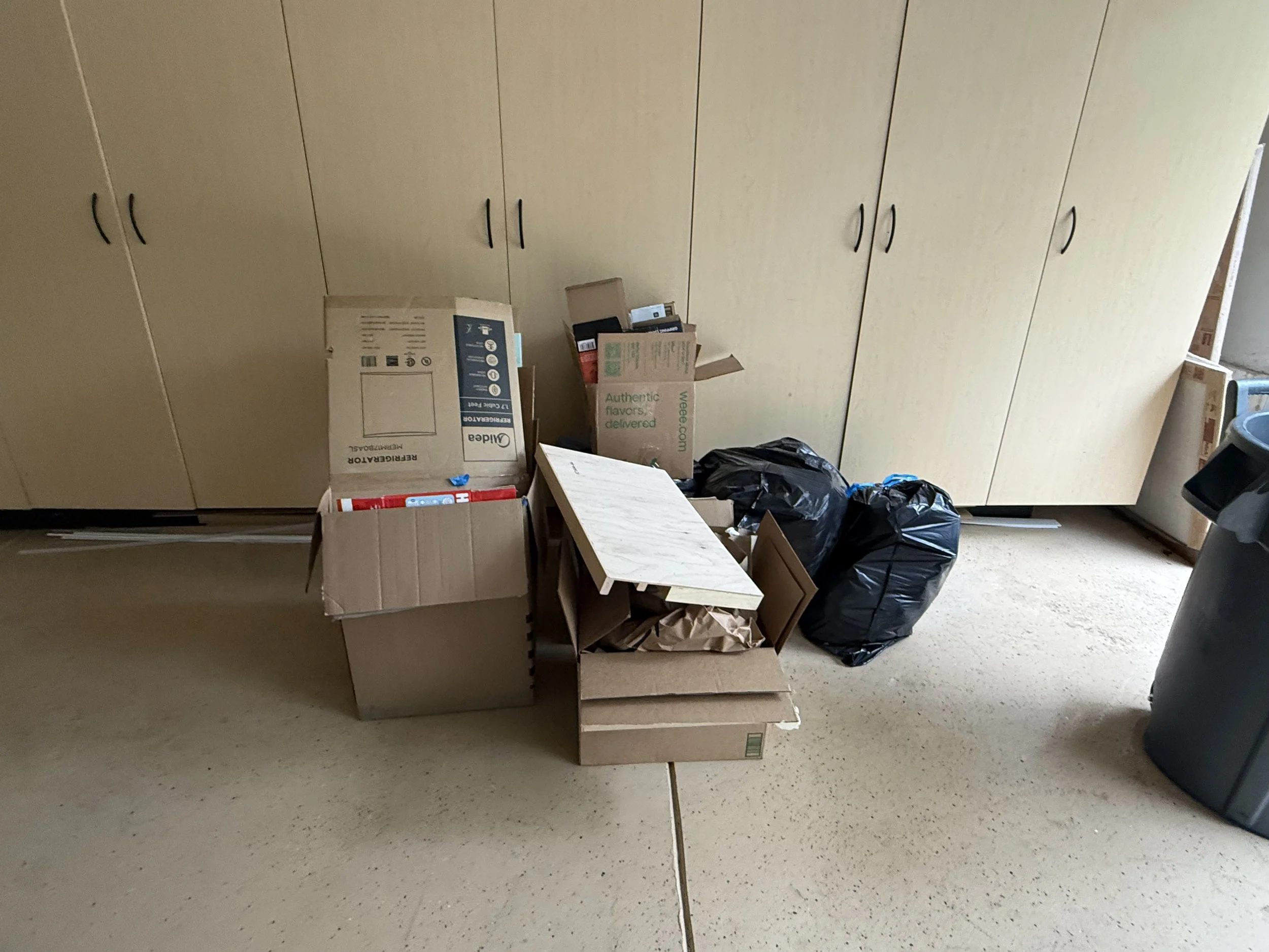 A room with beige storage cabinets, cardboard boxes, black trash bags, a small table, and various household items.