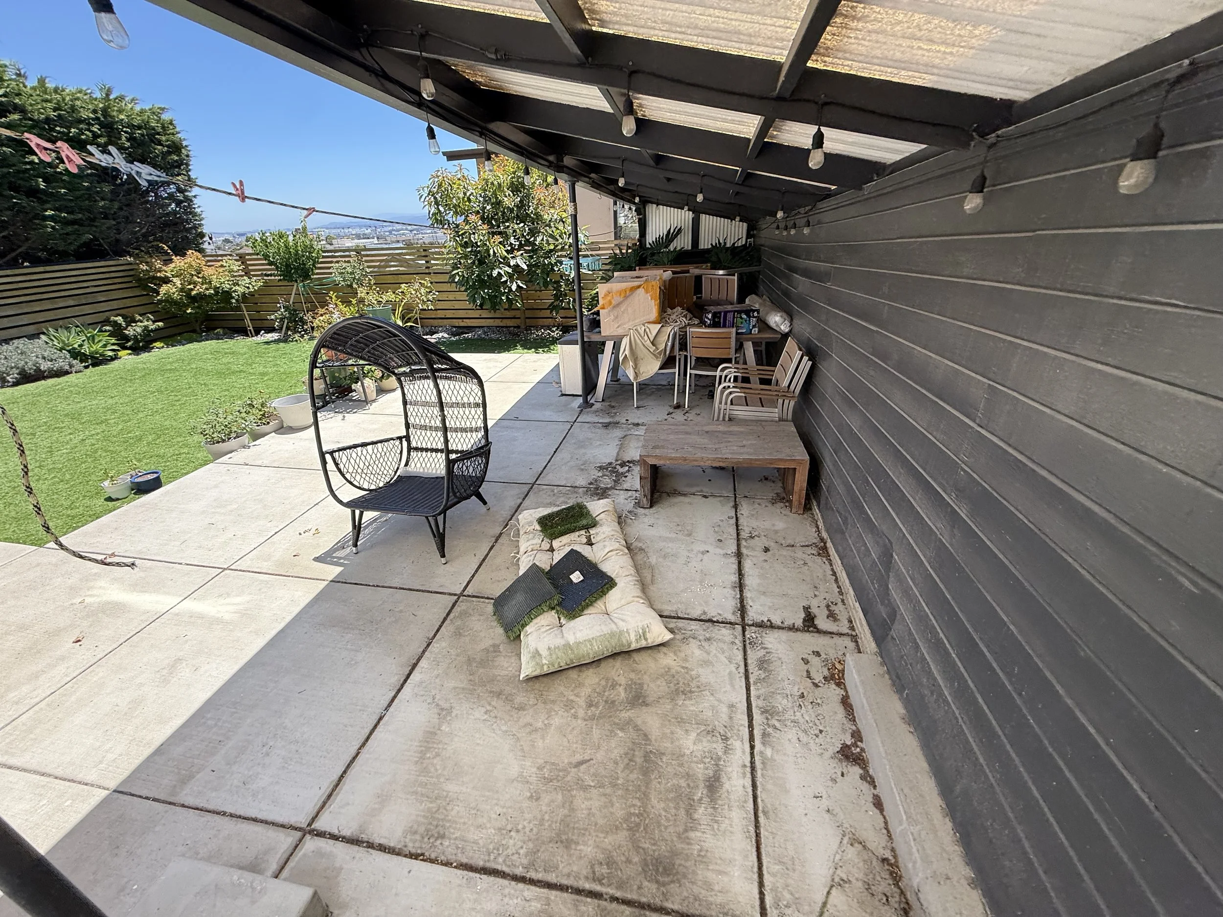 A backyard patio area with a black metal chair, a pile of pillows, a table with chairs, and various potted plants. There is a grassy area with a fence and trees in the background. String lights hang from the roof, which has a black and clear awning.