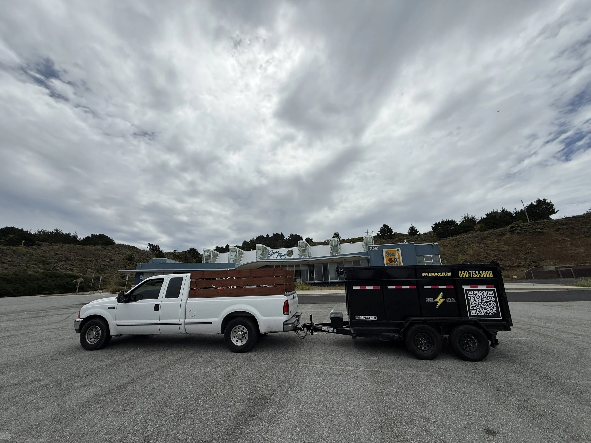 A white pickup truck towing a black trailer with a QR code on it, parked in an open lot with cloudy skies above and some trees and a building with a sign that says "Sea" in the background.