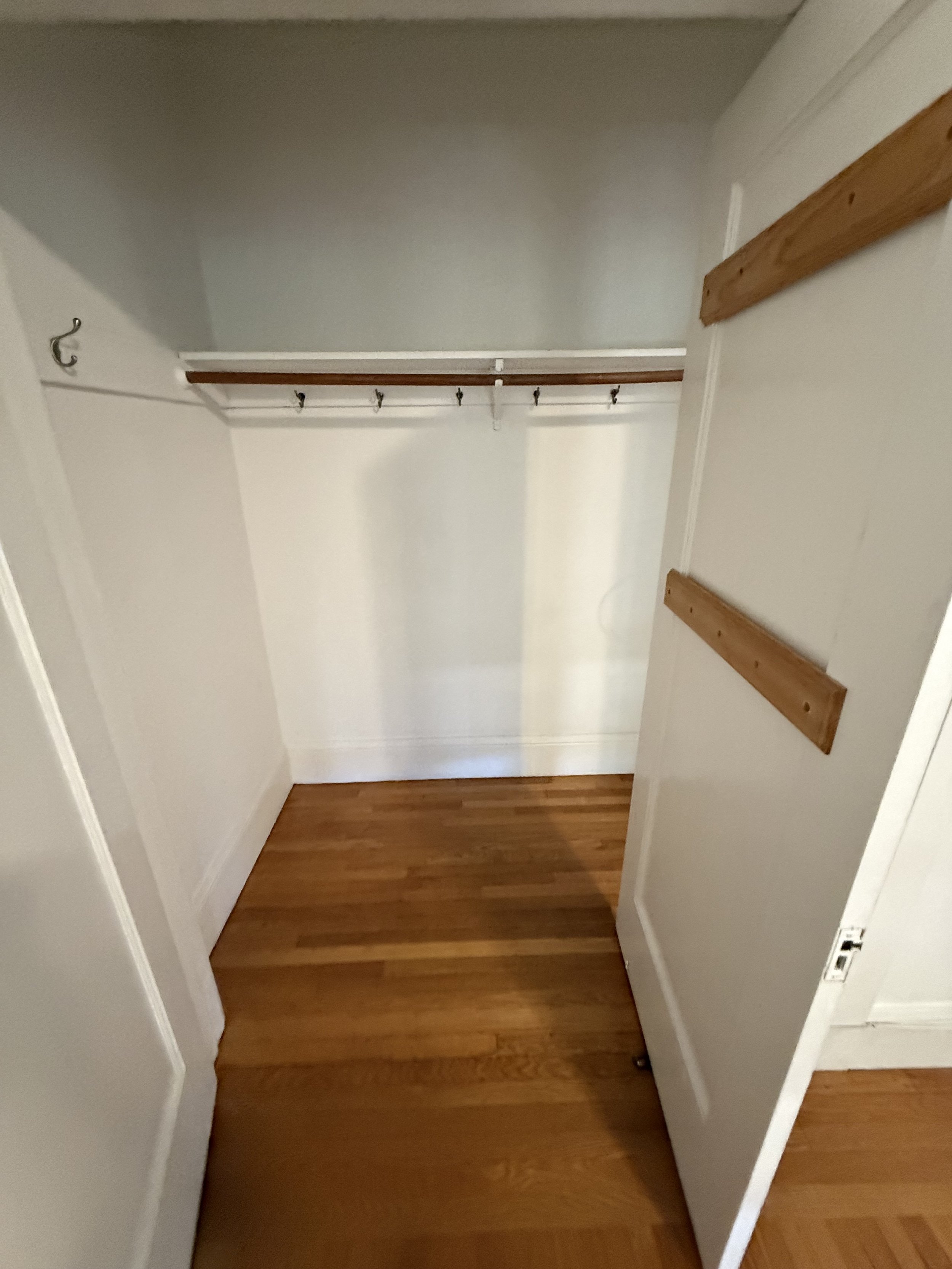 Empty closet with a wooden shelf and hooks, white walls, brown hardwood floor, and a white door frame.