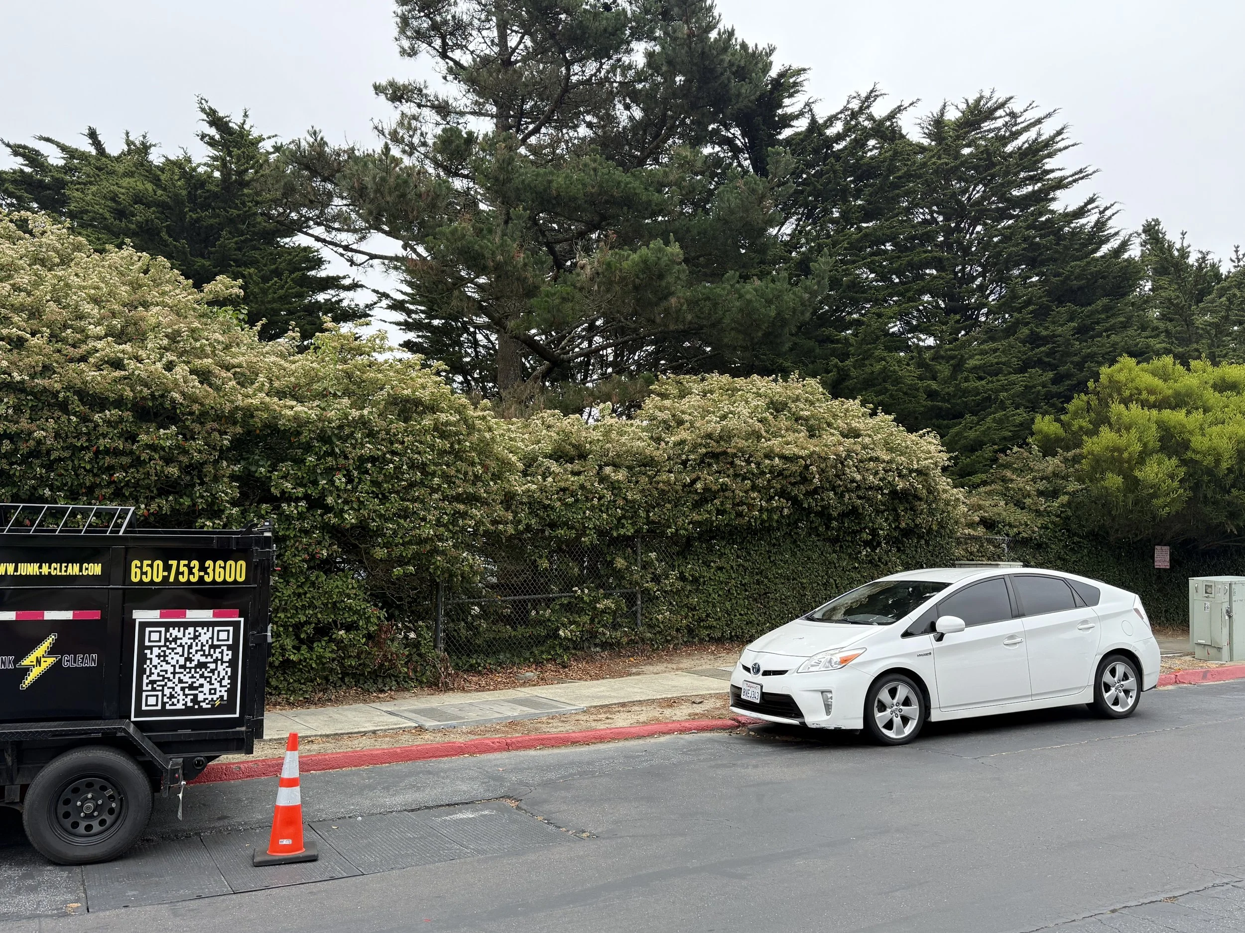 Parking lot with a white Toyota car parked, a black junk removal service trailer with a QR code and contact number, orange traffic cones, and green trees in the background.