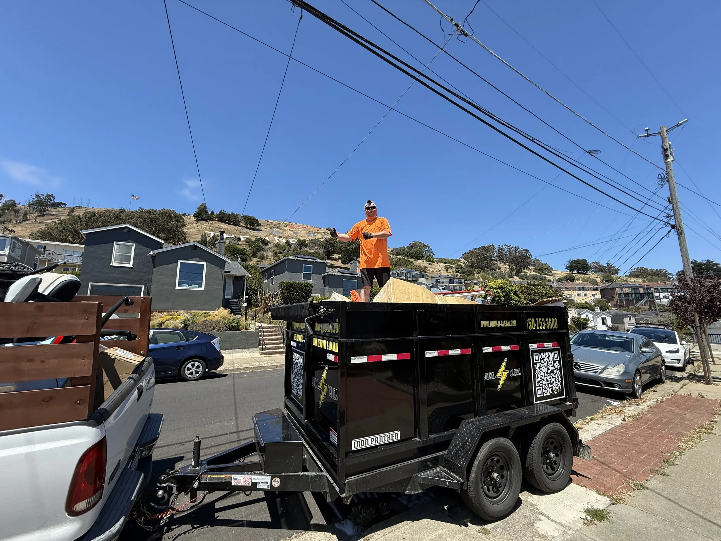 A man wearing sunglasses and an orange shirt standing on a trailer filled with debris, with a bright blue sky overhead. The trailer has logos and QR codes on the side, and there are residential buildings, parked cars, and power lines in the backgroun