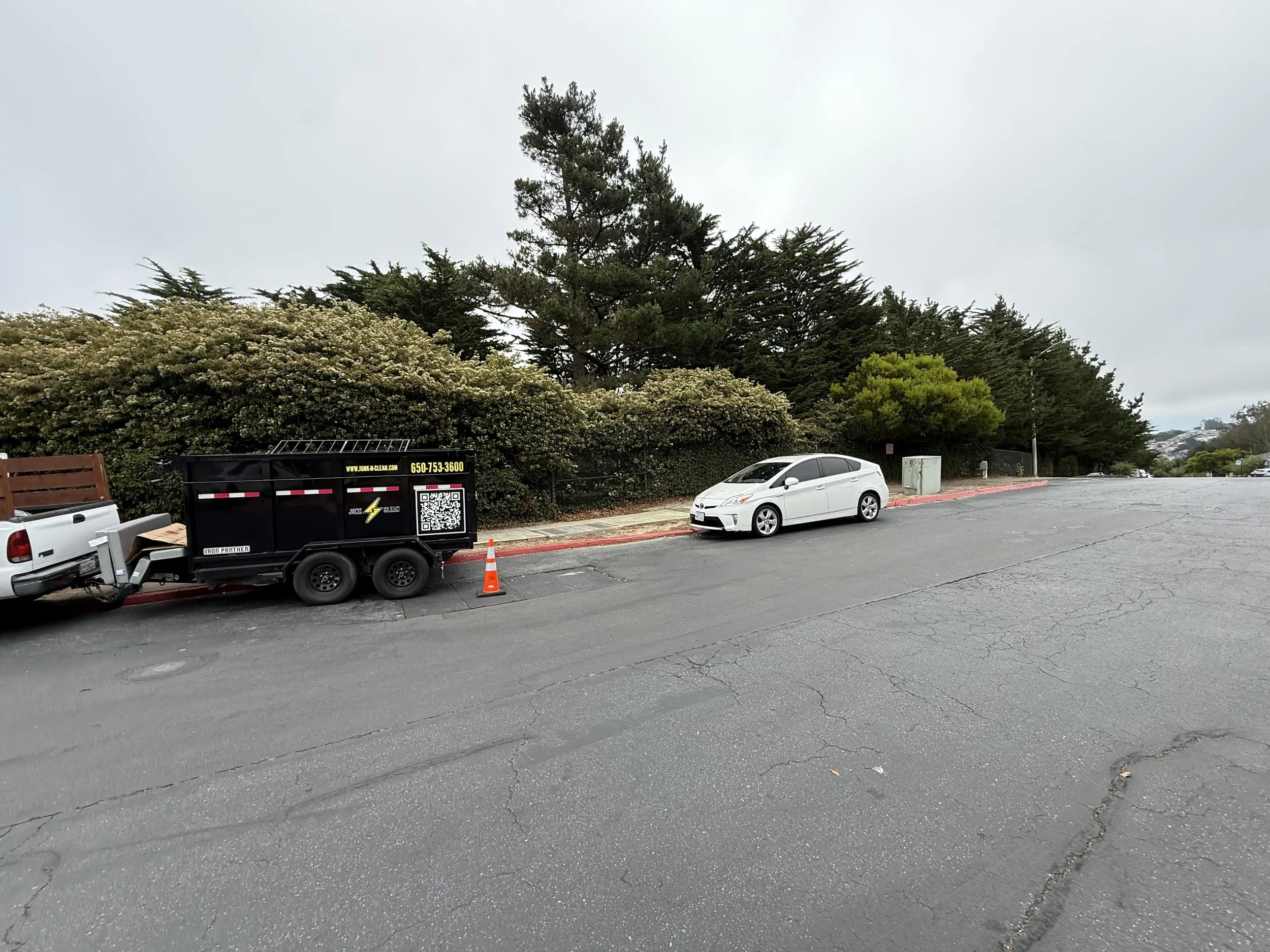 A parking lot with a white car parked along a curb, a black trash container, an orange safety cone, and a background of trees and cloudy sky.