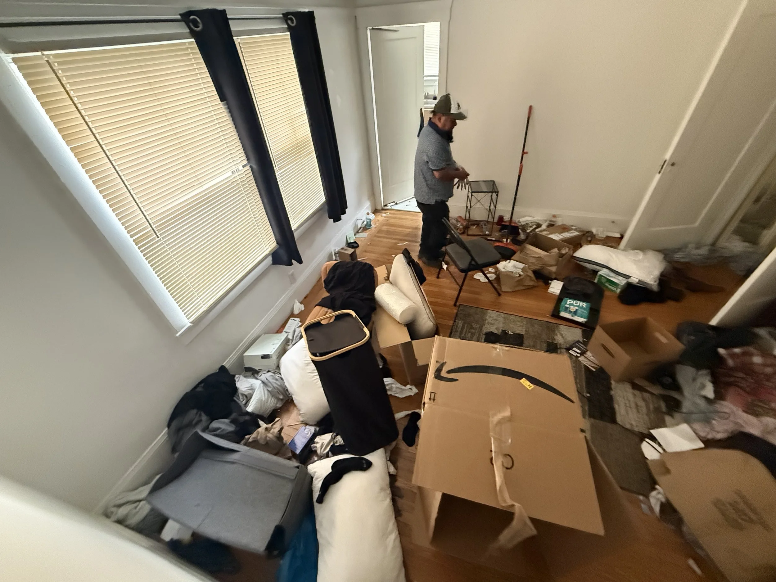 A man in a baseball cap and patterned shirt stands in a cluttered room next to a window with blinds. The room has a hardwood floor and various boxes, bags, and miscellaneous items scattered around.