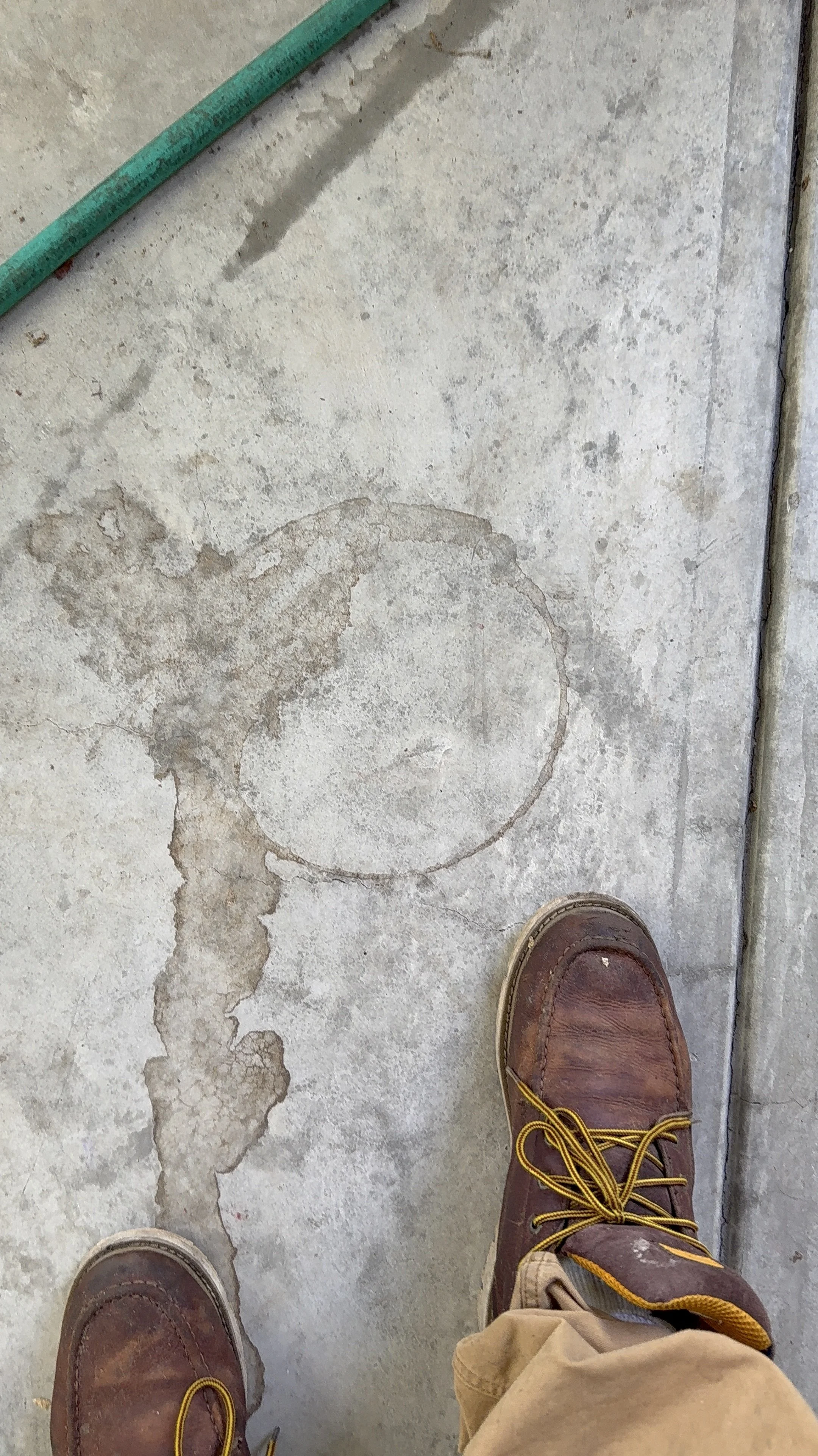 A person's feet wearing brown work boots with yellow laces are standing on a concrete floor with dark stains and a faint circular outline. Part of the person's beige pants is visible. A green measuring tape and a small twig are on the floor.