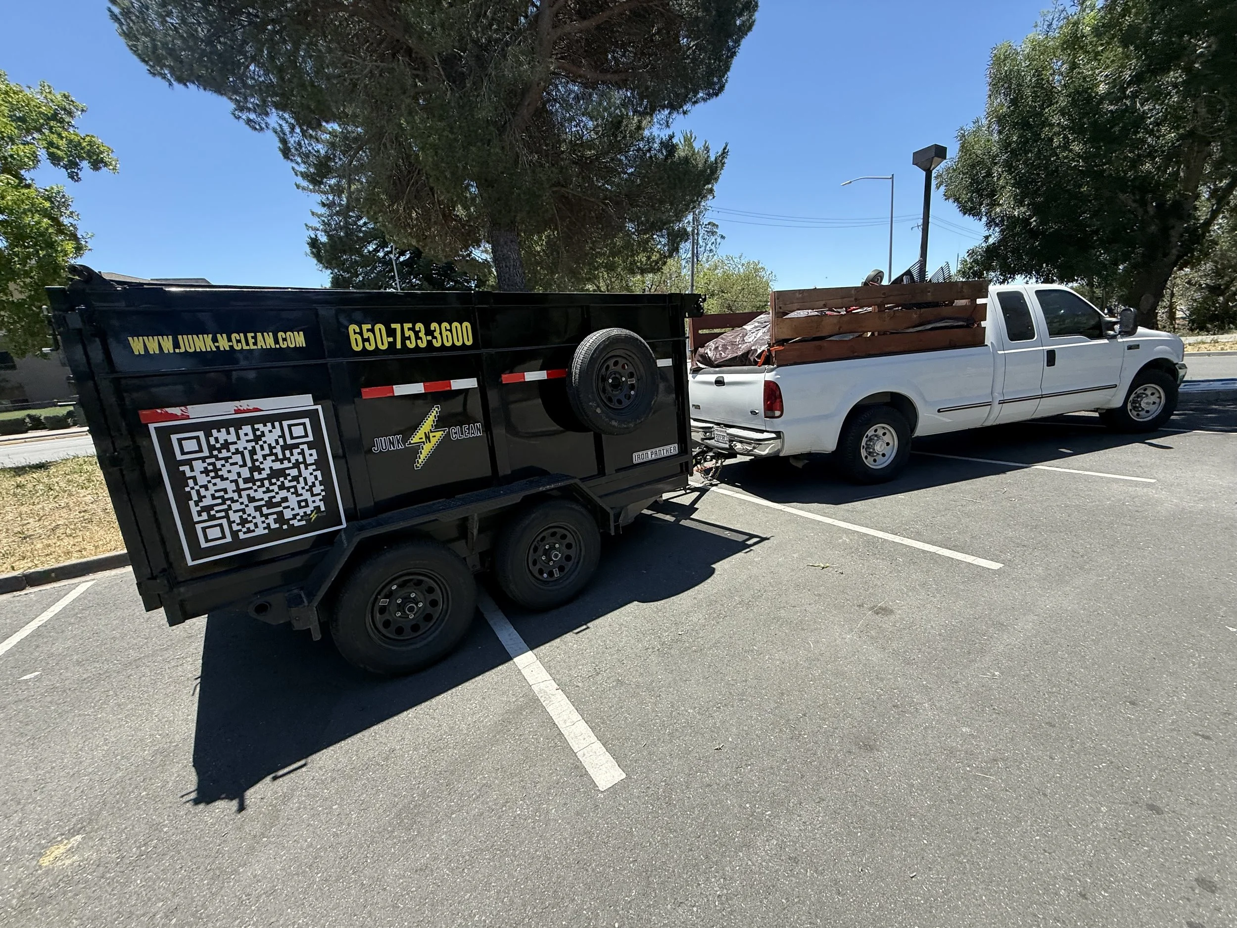 A white pickup truck and a black trailer parked in a parking lot under tall trees with green leaves and a clear blue sky.