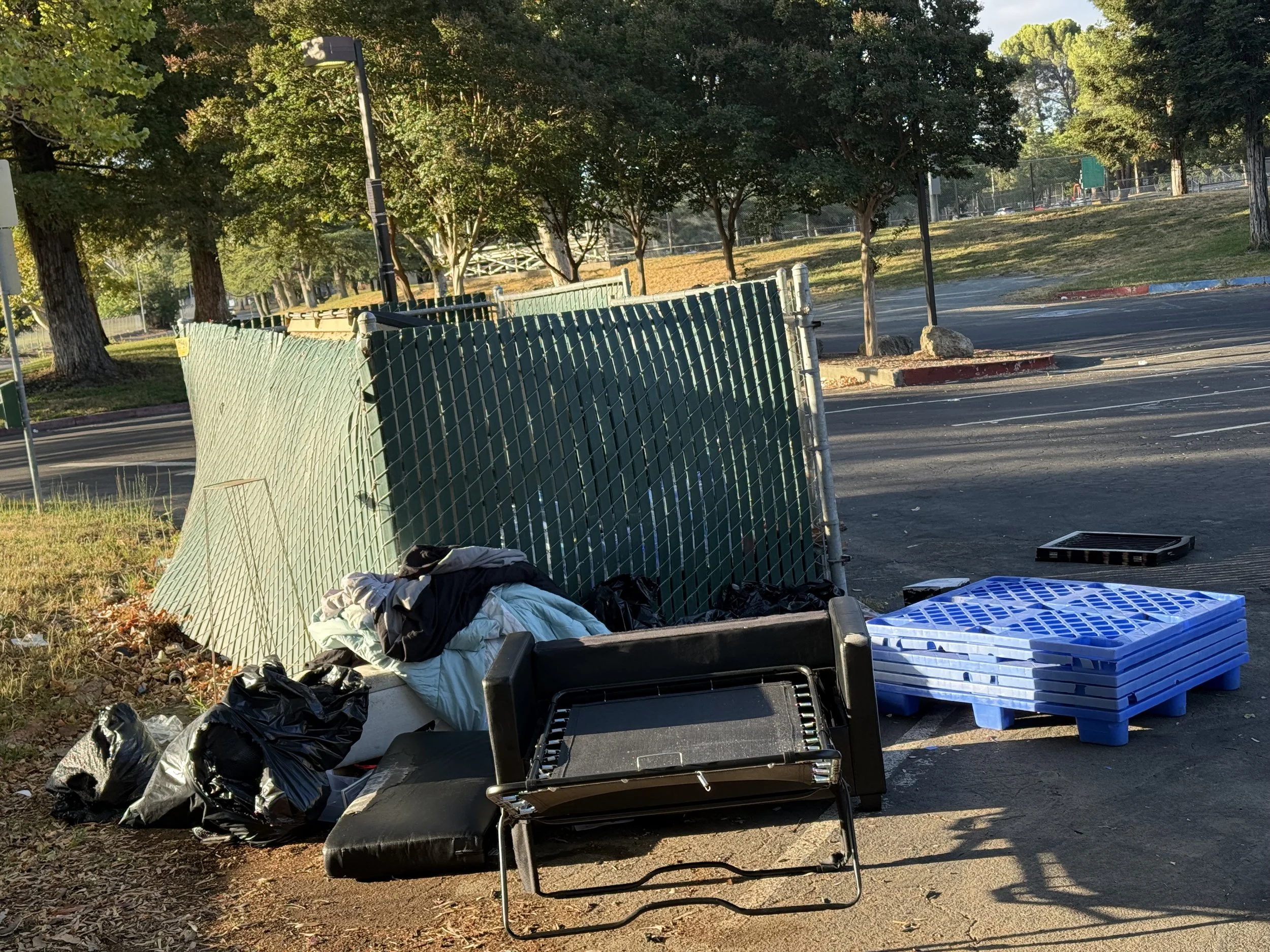A fenced-off construction or cleanup site with scattered black trash bags, plastic pallets, and various debris on the ground, with trees and a park in the background.