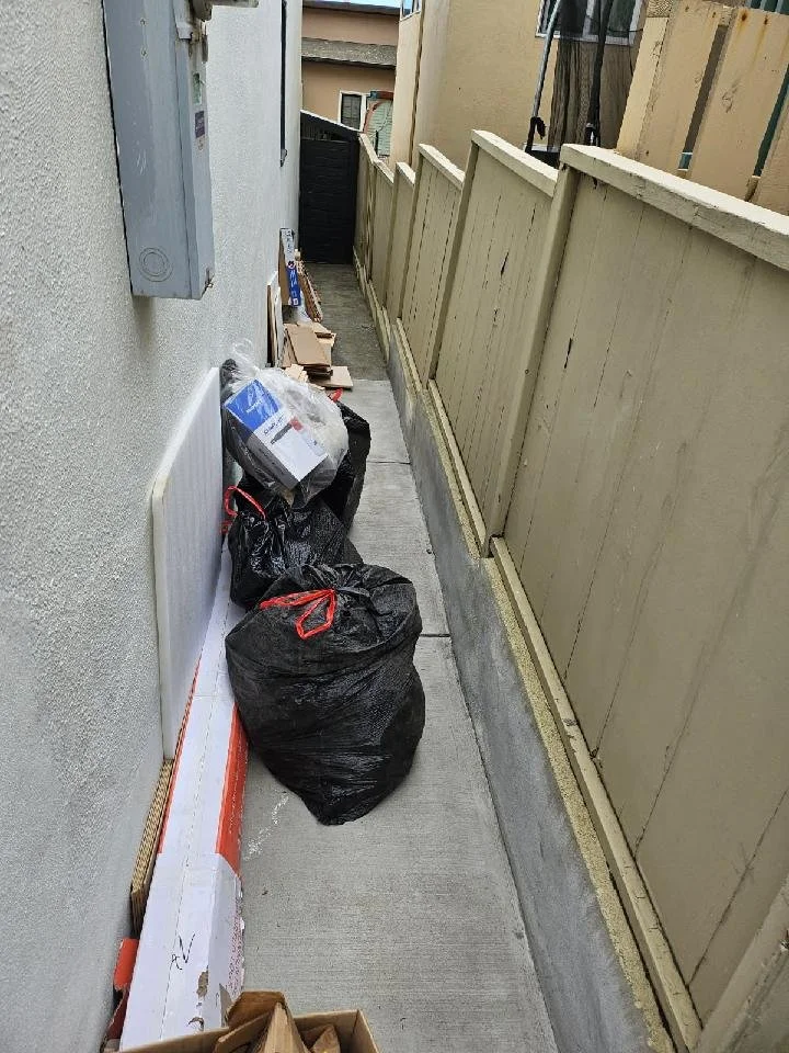 Small outdoor alleyway with bags of trash, cardboard boxes, and scattered debris along a concrete pathway with a beige wooden fence on one side and a house wall on the other.