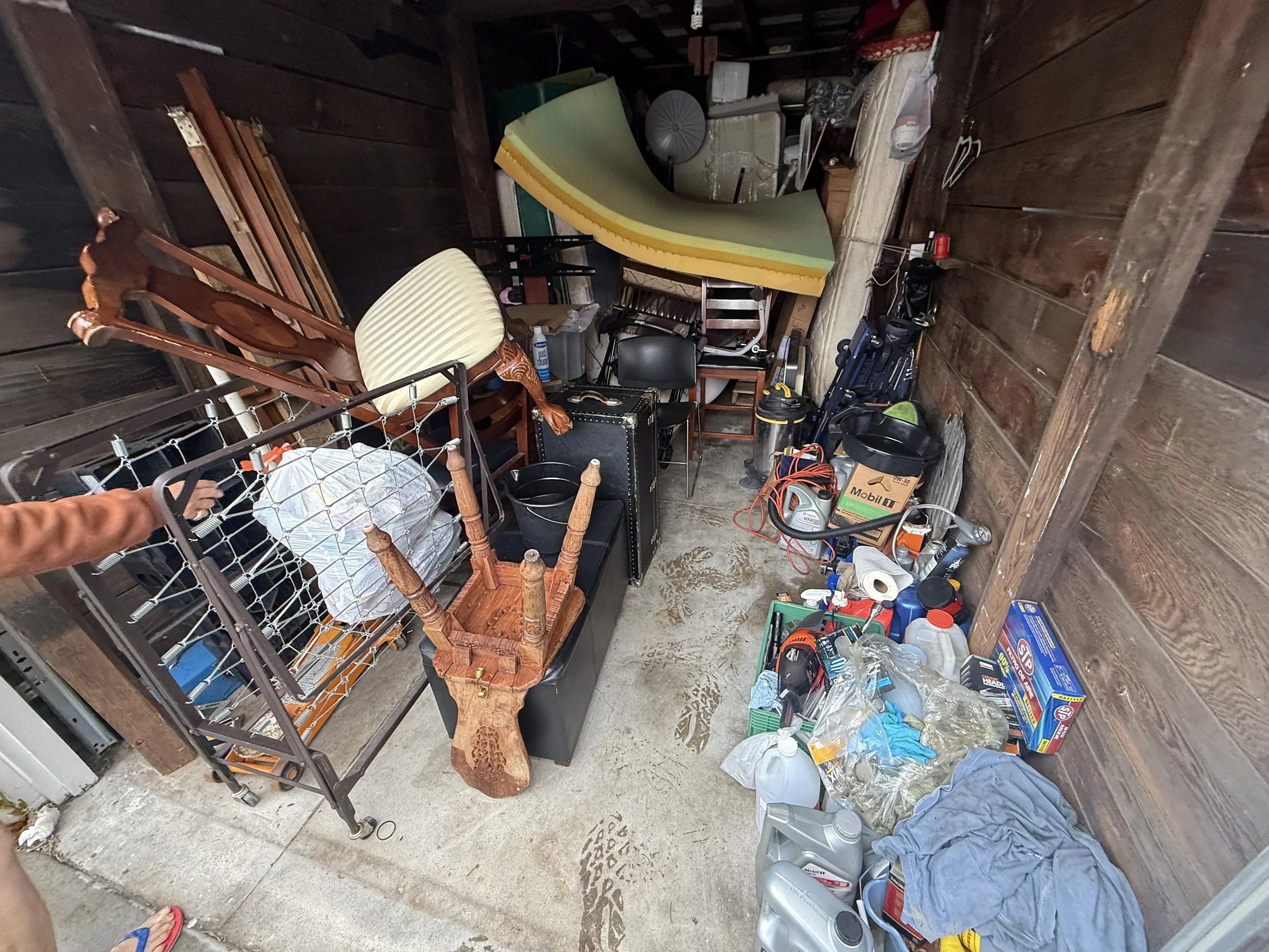 A cluttered garage with various household and tool items, including chairs, a foam mattress, storage boxes, vacuum cleaner, and trash bags, on a concrete floor with visible footprints.