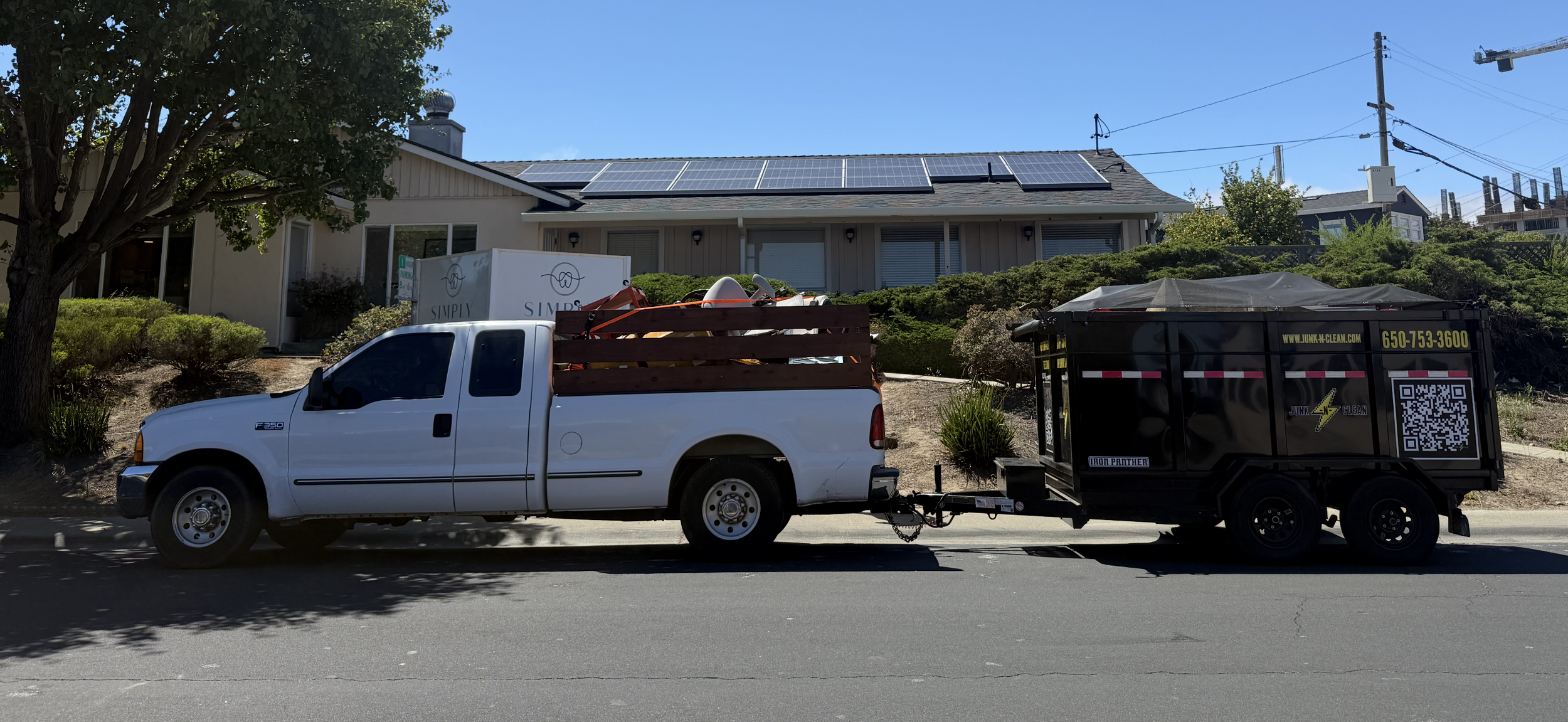 A white pickup truck with a wooden frame carrying items, connected to a black junk removal trailer with yellow and white text, parked in front of a house with solar panels on the roof and a tree on the left.