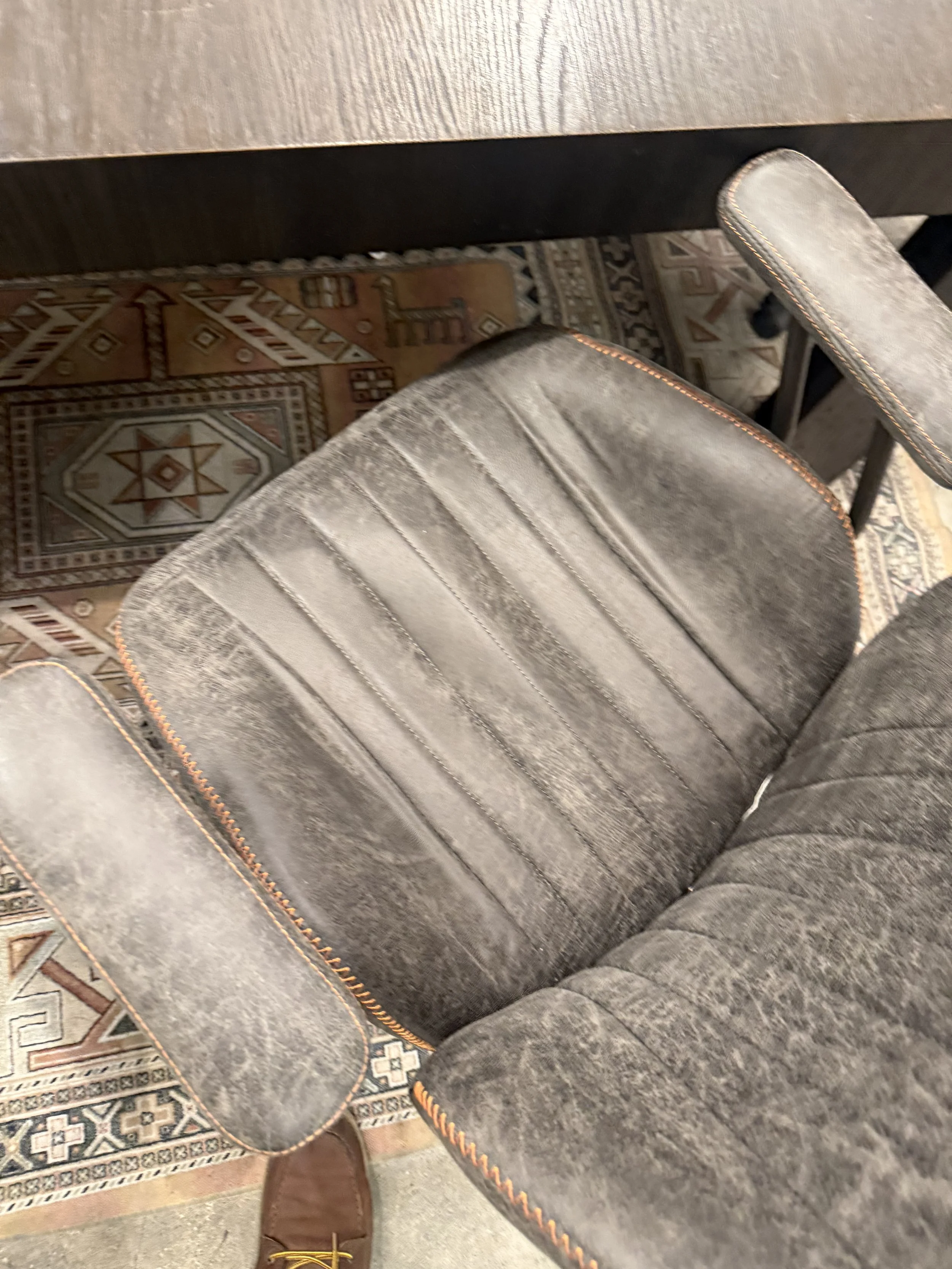 A person's foot in a brown shoe stepping onto a patterned rug next to a gray upholstered chair with orange stitching on a wooden floor.