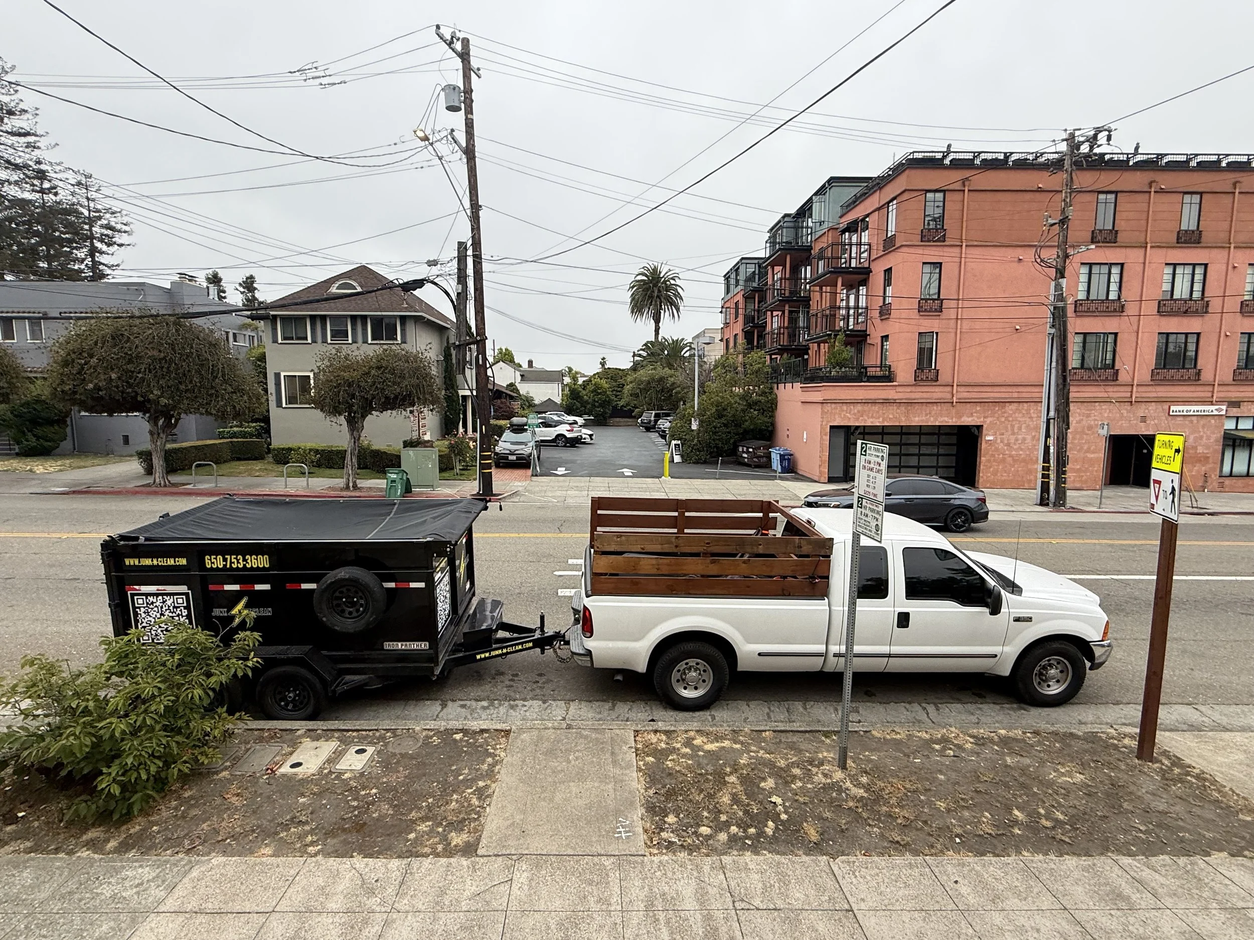 City street scene with commercial and residential buildings, parked cars, utility poles, and overcast sky.