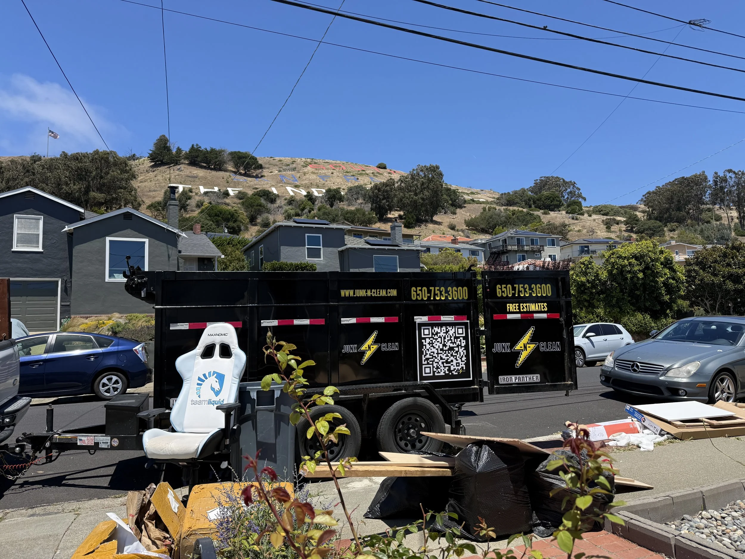A backyard with a hill featuring large painted letters that spell 'FREMONT.' Several houses are visible in the foreground, along with plants, trees, and a utility pole with wires. There are various items on the driveway, including a black trailer wit