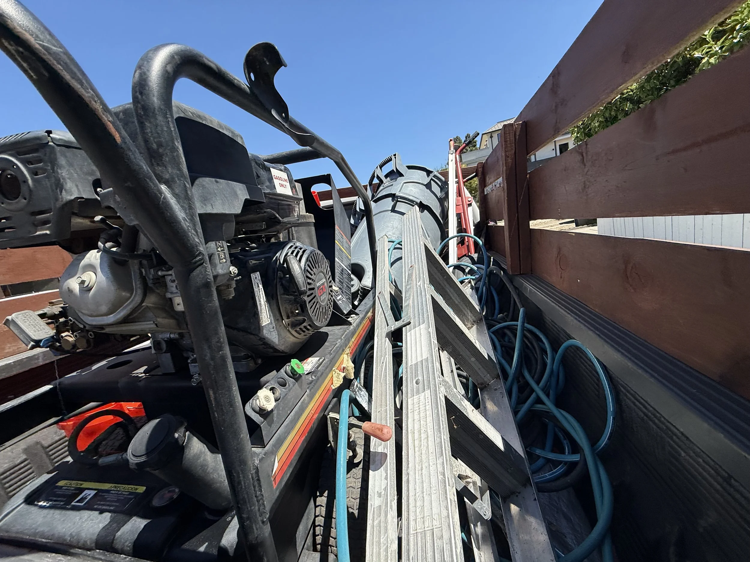 The image shows the bed of a pickup truck loaded with various tools and equipment, including a gas-powered generator, metal ladders, extension cords, and other maintenance supplies, under a clear blue sky.