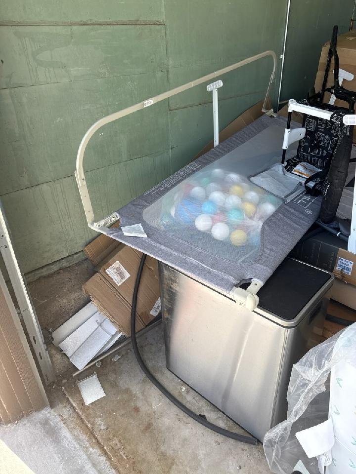 A baby changing station with a transparent cover, containing colorful balls, positioned above a metal trash can in a storage room with cardboard boxes and miscellaneous items around.