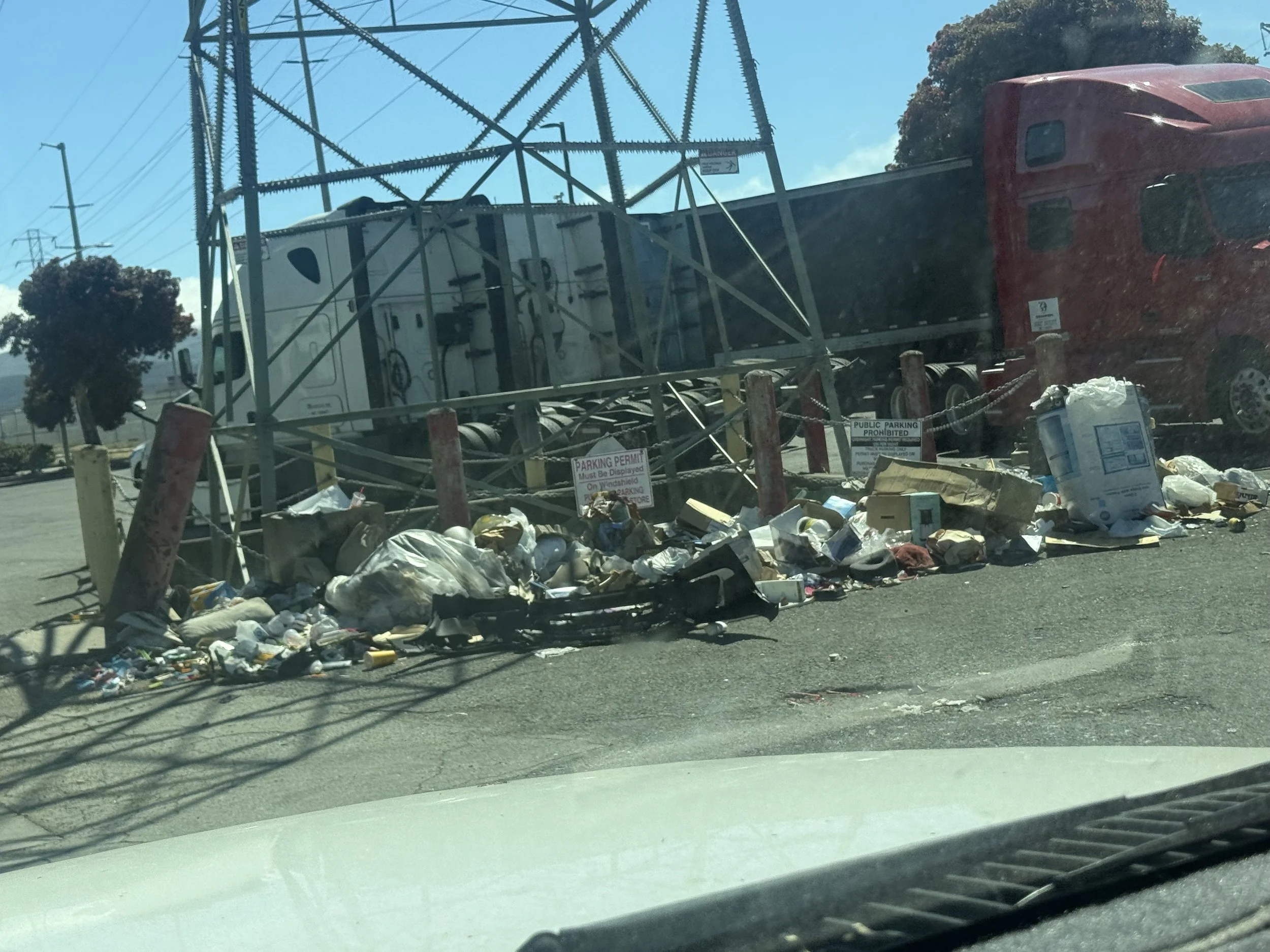 A red semi-truck is overturned near a utility pole and power lines, with debris and trash scattered around on the ground in a parking lot or roadside area.