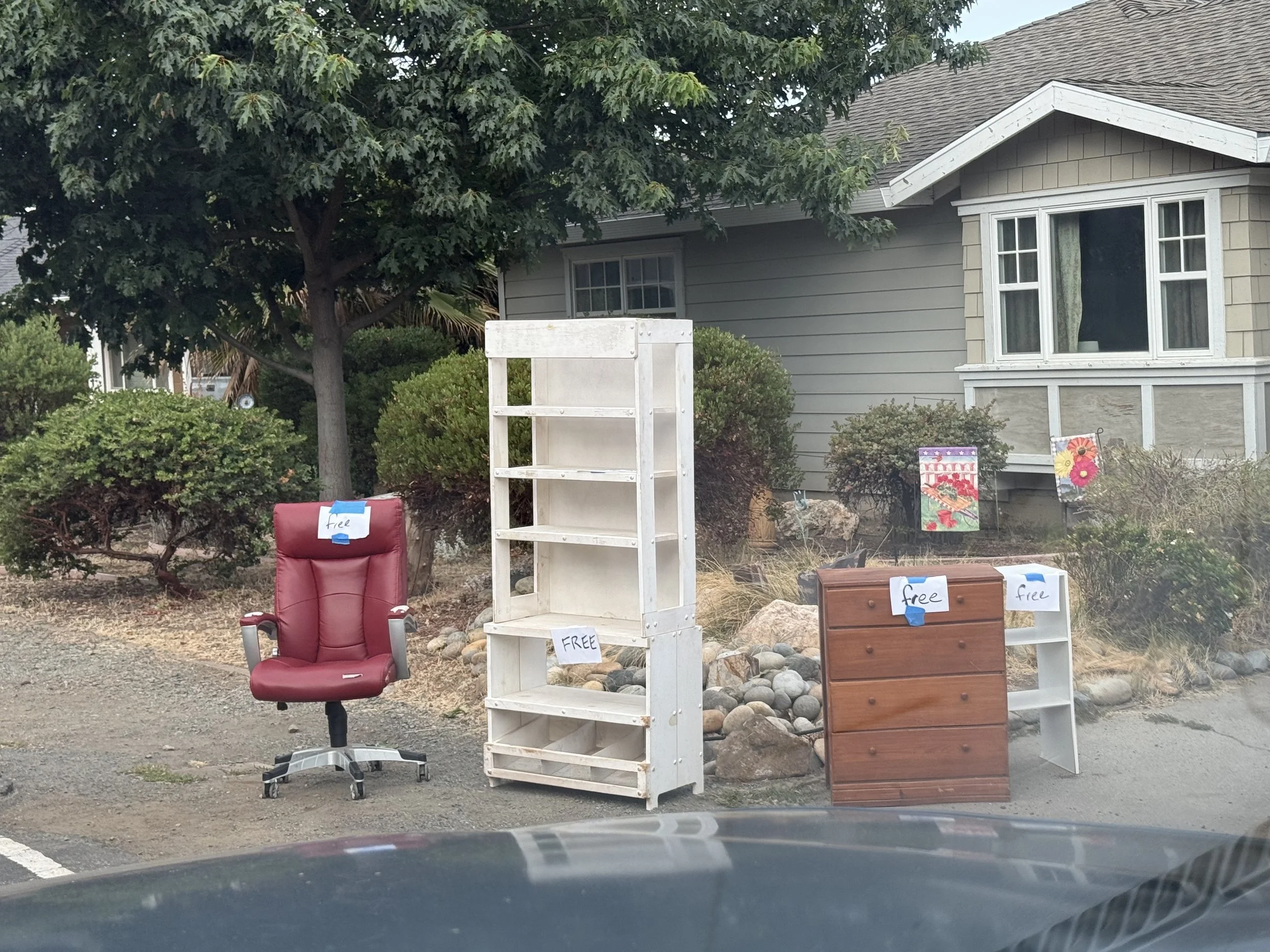 A yard sale setup with a red office chair and empty white shelves with a free sign, brown wooden drawers, and small posters with flowers. There is a house with gray siding, a bay window, and a large tree in the background.