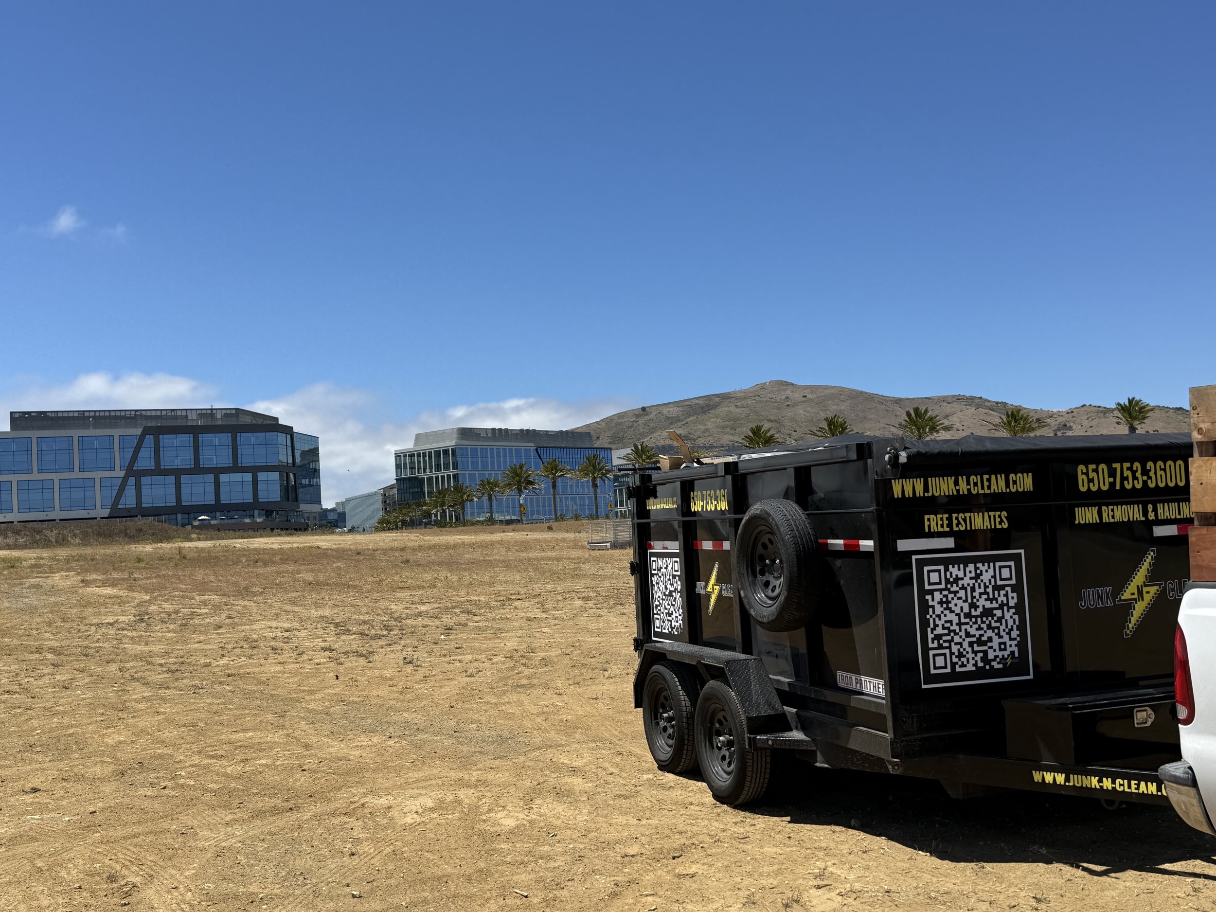 A cityscape with modern glass buildings, palm trees, a hillside, and a pickup truck with a black trailer attached parked on a dirt field under a partly cloudy blue sky.