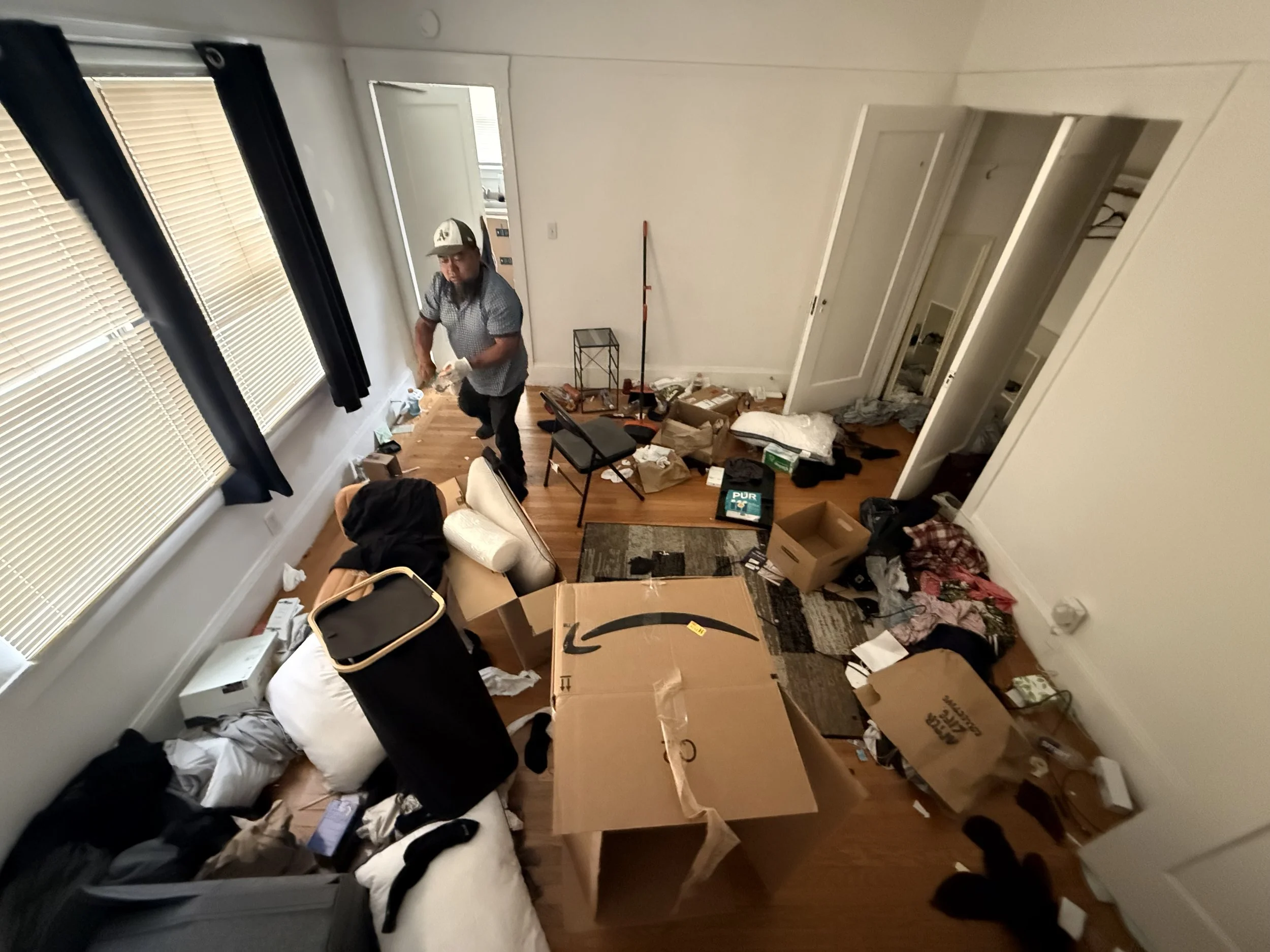 A person standing in a cluttered room with boxes, clothes, and furniture.