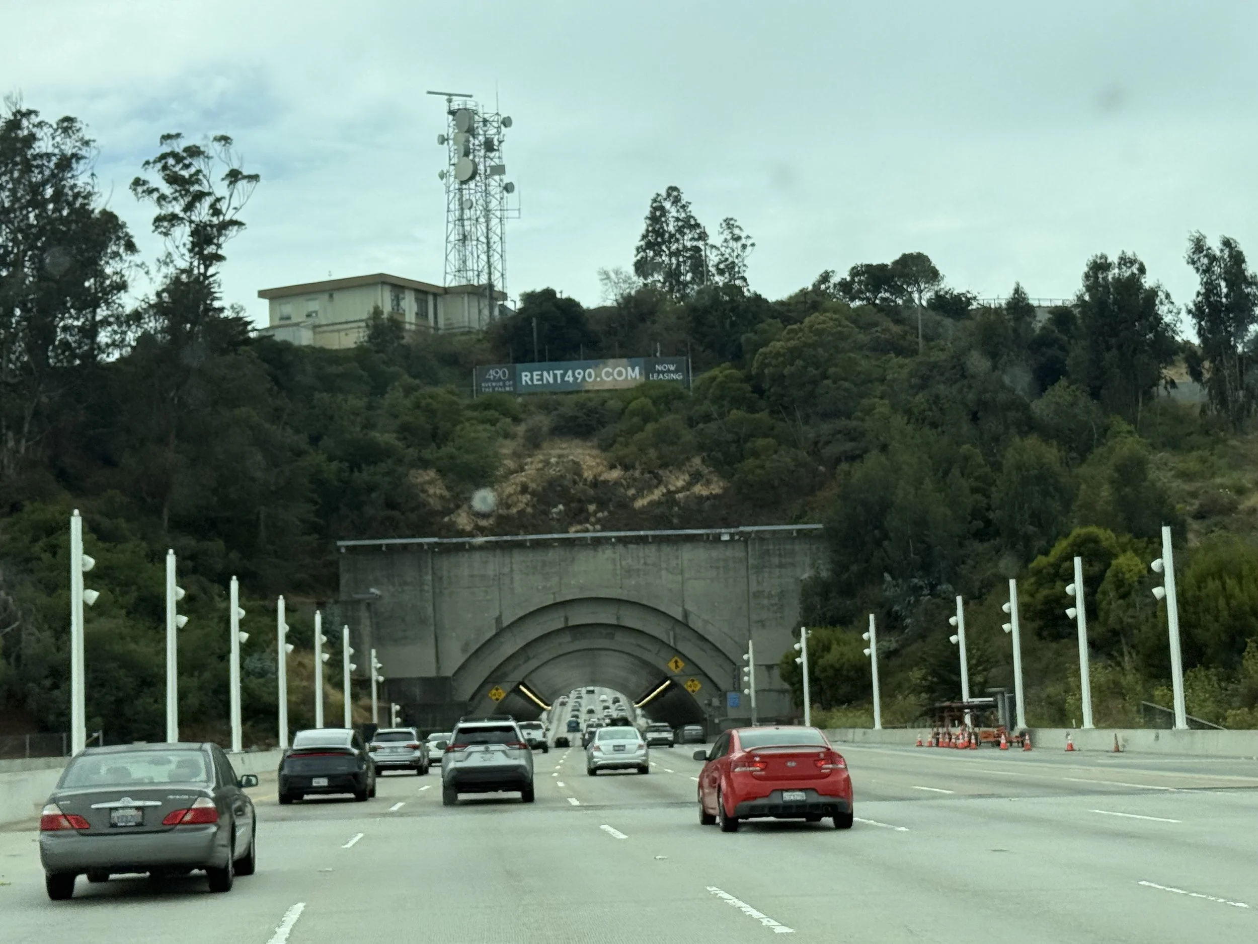 View of a highway with multiple cars driving under a tunnel carved into a hillside. In the background, there is a communication tower with antennas and satellite dishes on a hilltop, as well as a building. A billboard advertising rental properties is