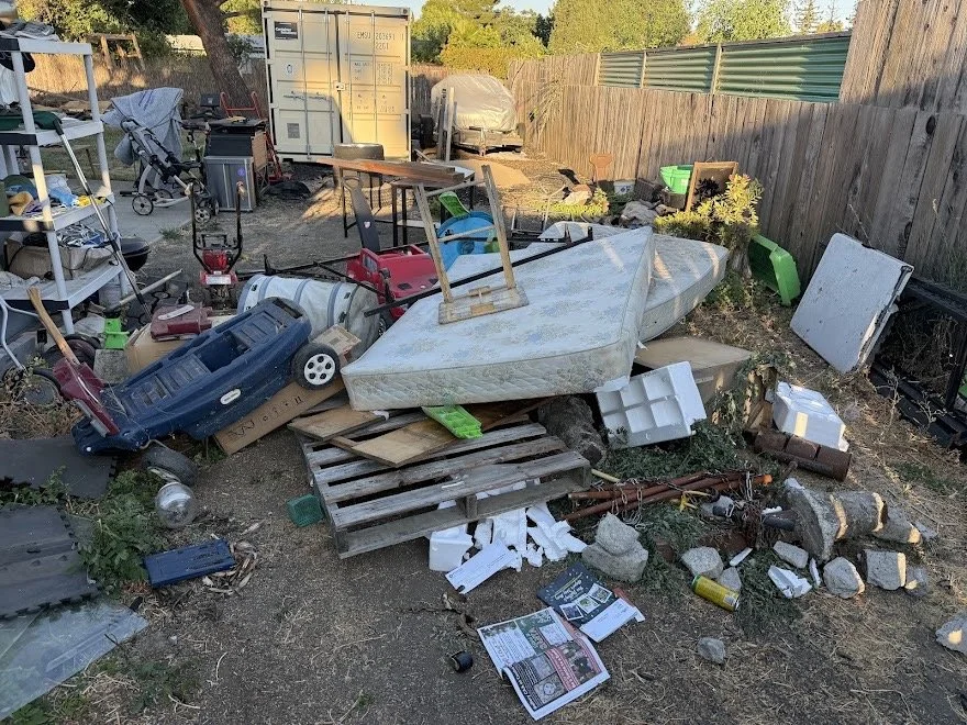 Disorganized backyard with a small boat, various children's toys, tools, and miscellaneous items scattered on the ground, surrounded by a wooden fence and storage containers.