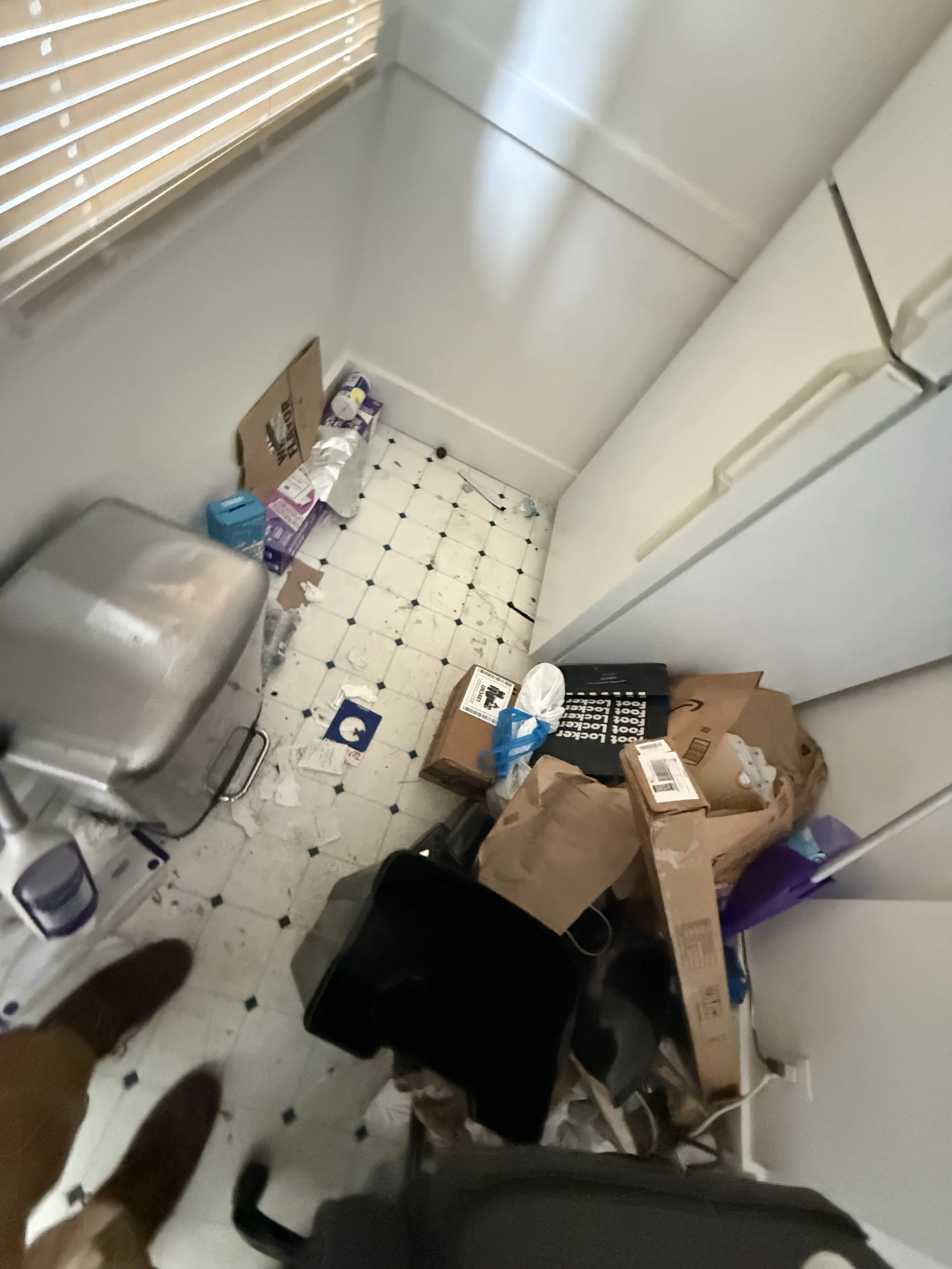 A cluttered small room or pantry with various boxes, bags, and items scattered on the floor. There is a white appliance, possibly a microwave or toaster oven, and a white wall with a radiator on the left. The floor is covered with white tiles with bl