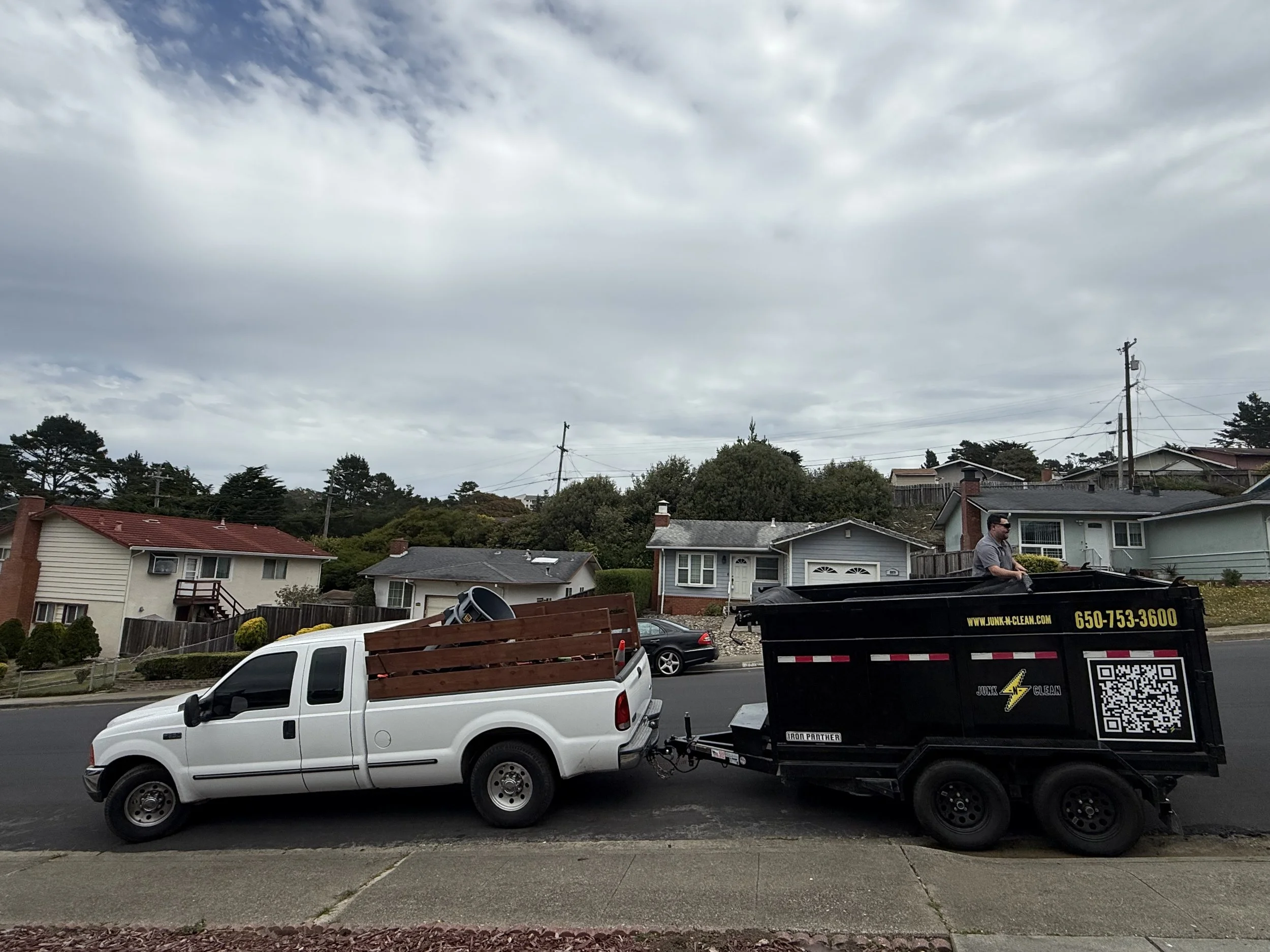 Residential neighborhood street with houses, trees, and utility poles underneath a cloudy sky. A white pickup truck is parked, hauling a box trailer with a man standing next to it, near a black dumpster.