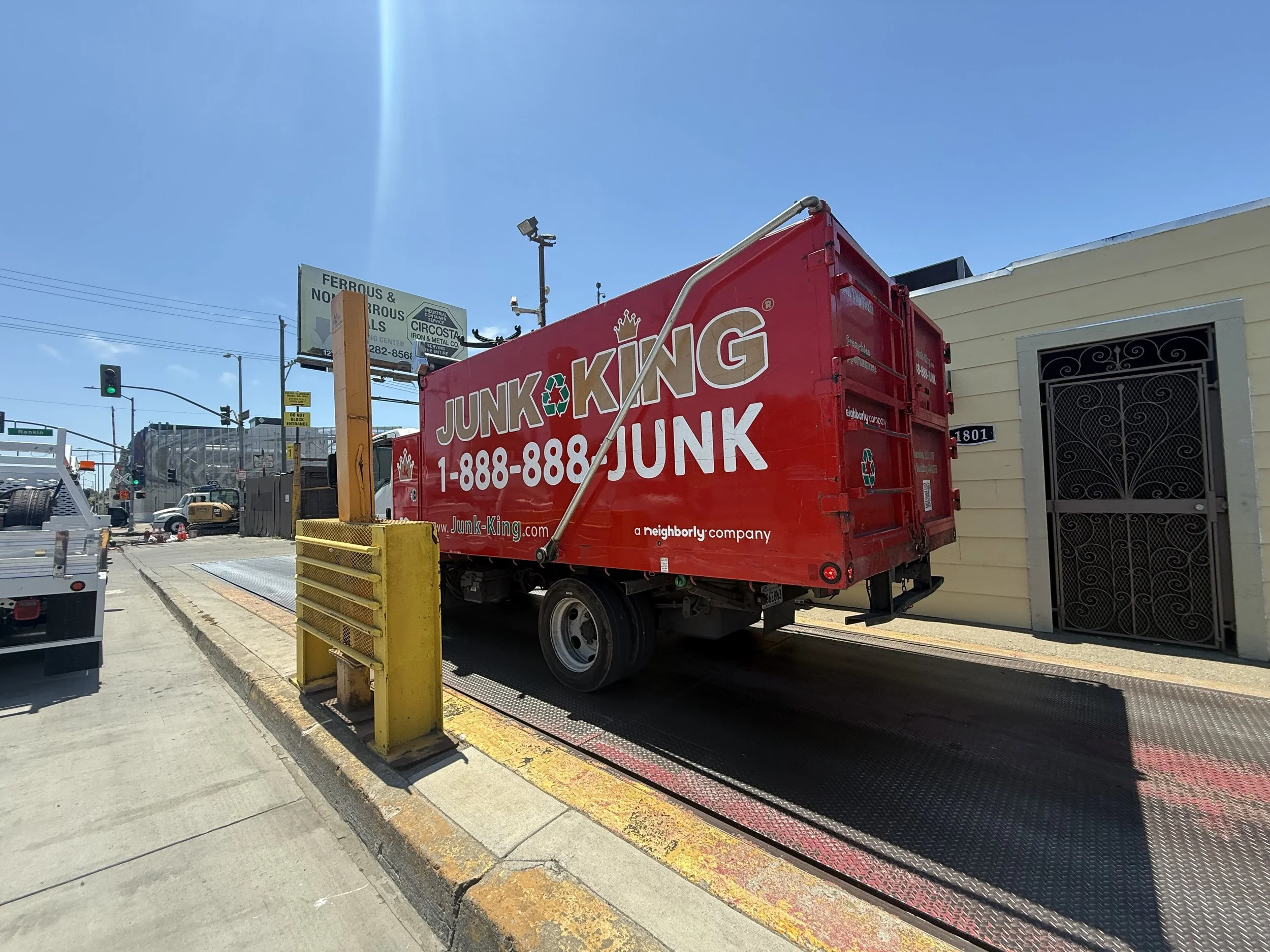A red junk removal truck parked on the street near a yellow building, with a sign for industrial services and a traffic light in the background.