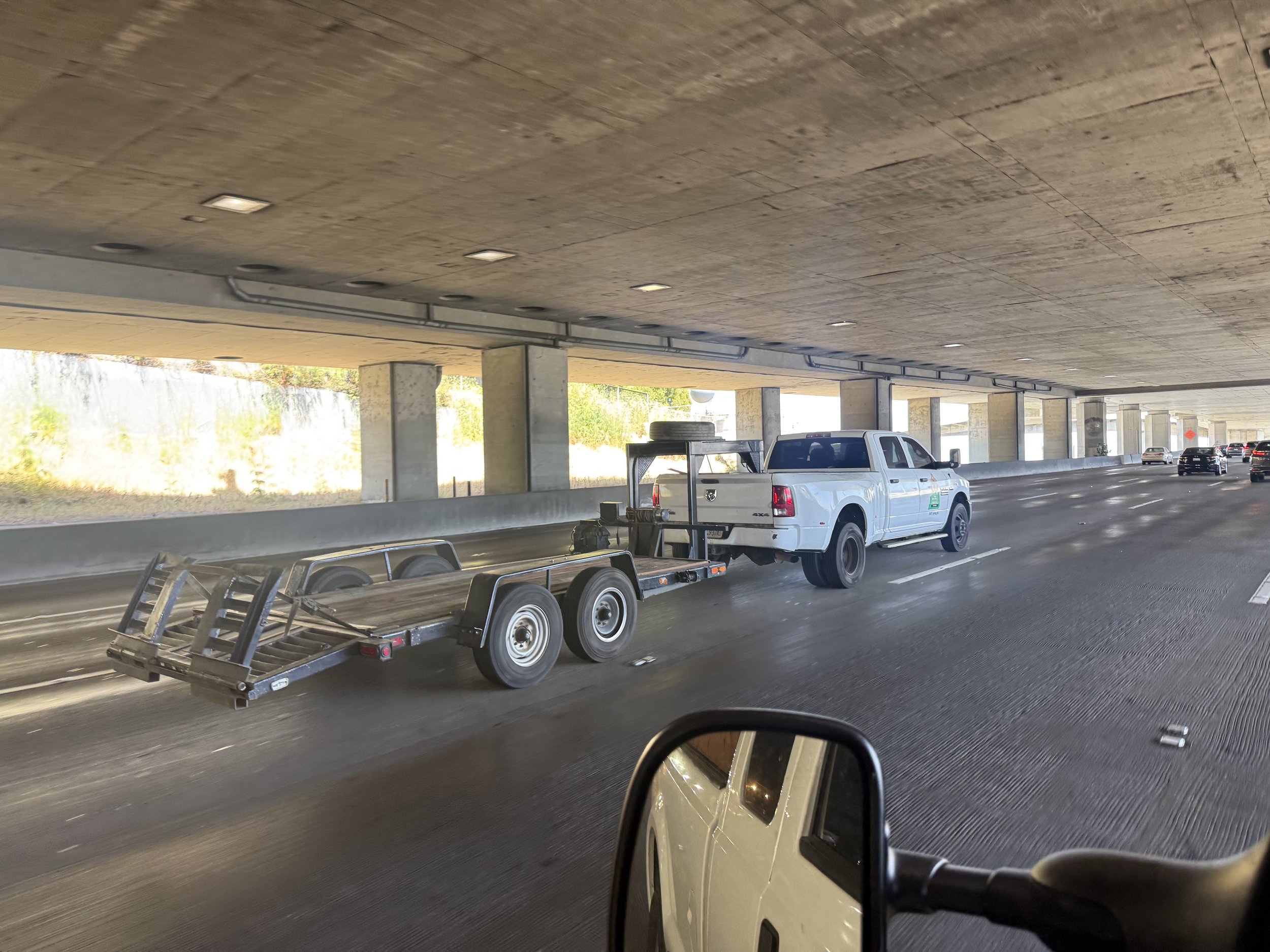 Underneath a concrete highway overpass, there is a white pickup truck towing a empty flatbed trailer, with other vehicles and a wall in the background.