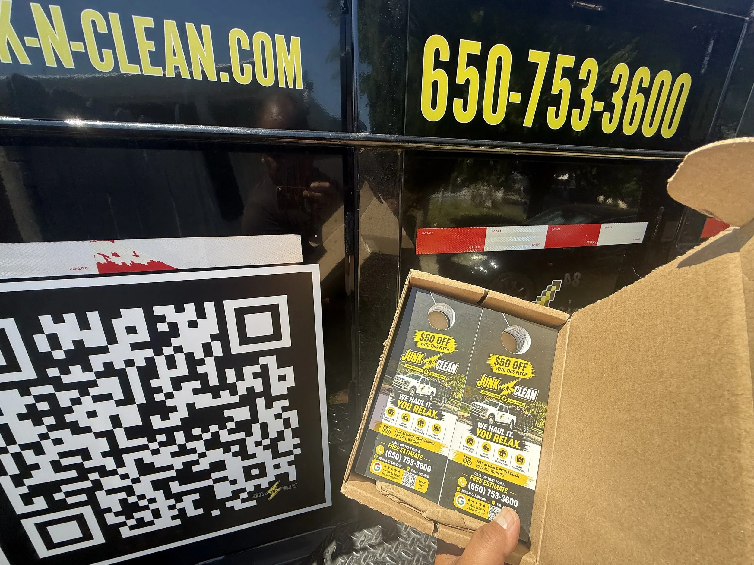 A person holding a flyer for a junk removal service called 'Junk N Clean' with a photo of a truck, offering $50 off with the flyer, and contact information. Nearby is a large QR code sign and a part of a customer receipt.