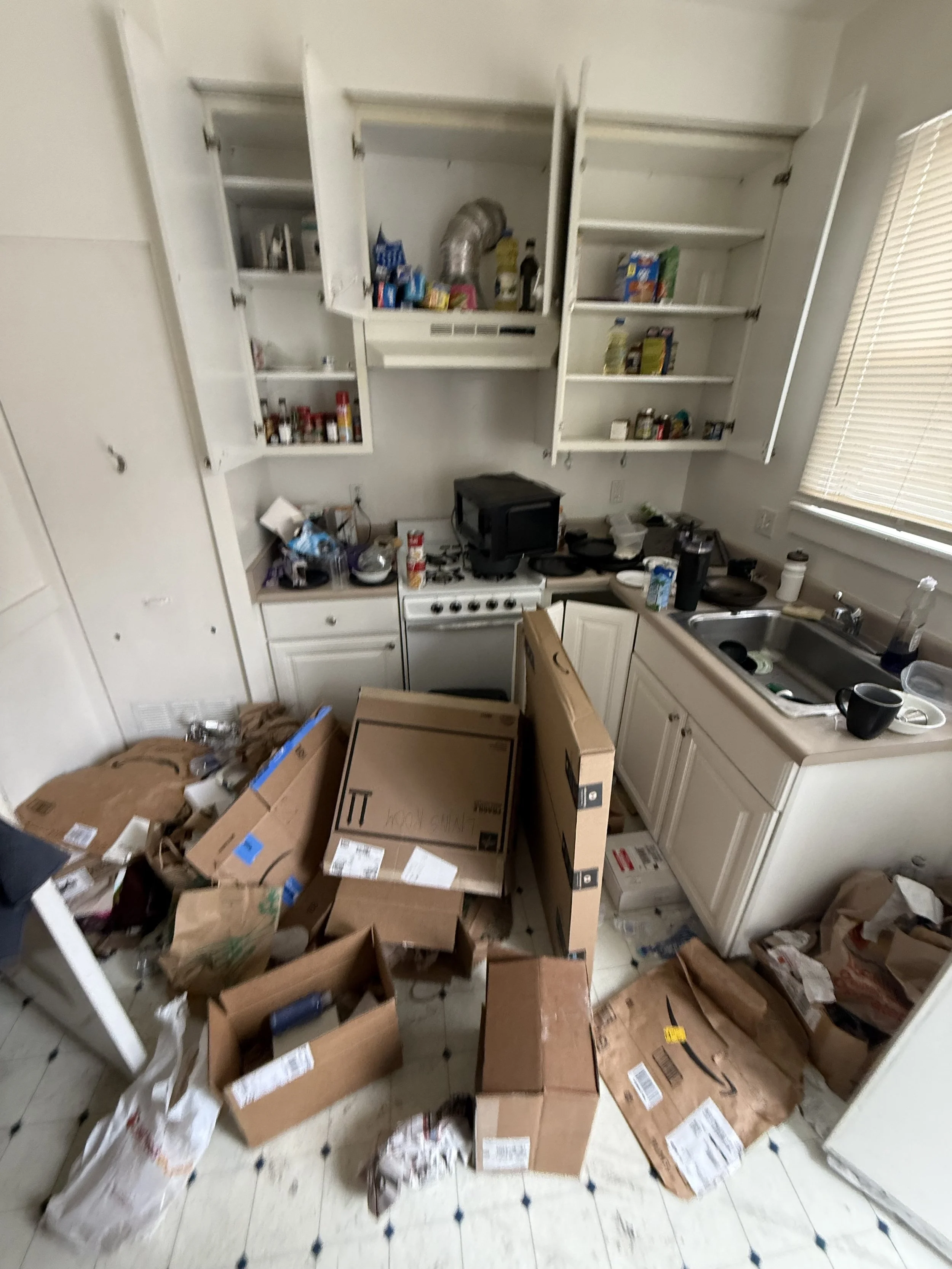 Cluttered kitchen with open cabinets, boxes, and various items on the counter and floor