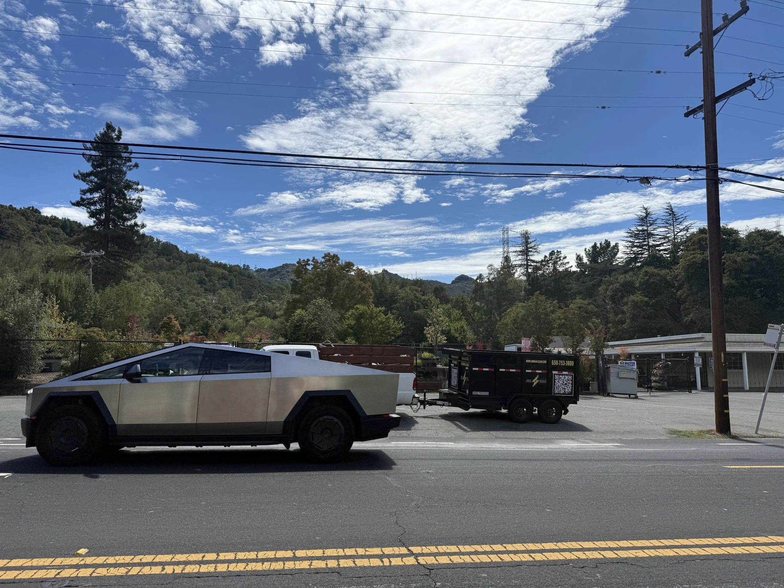 A Tesla electric pickup truck parked on a road with a mountain and blue sky with clouds in the background, utility poles, a trailer, and trees surrounding the area.