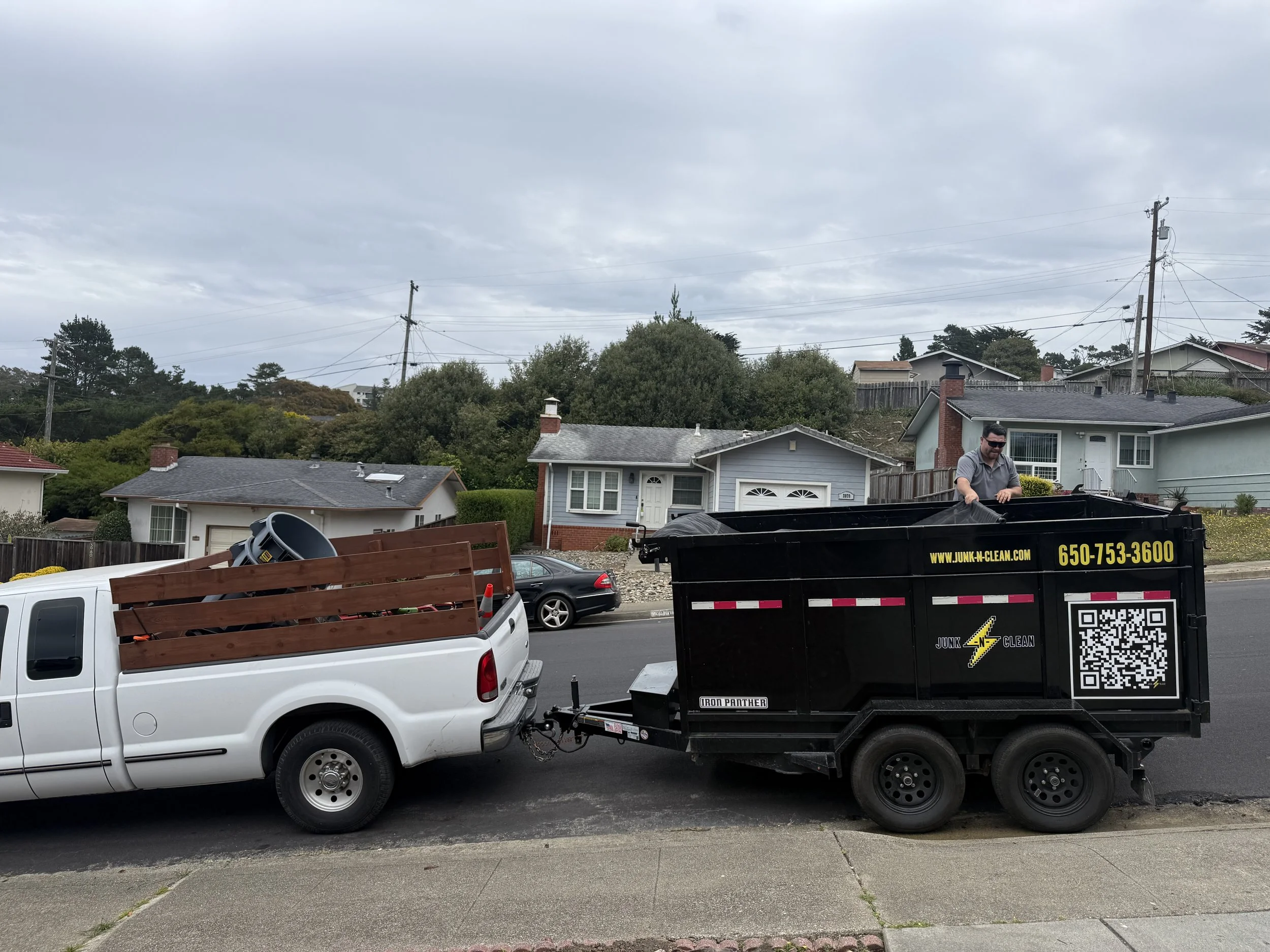 A man standing in the back of a black junk removal truck on a residential street, with houses and trees in the background, on a cloudy day.
