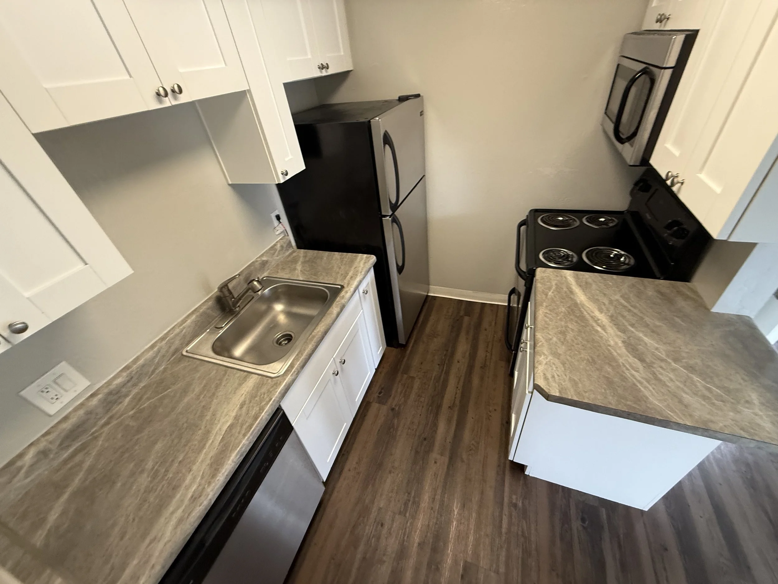 A compact kitchen with white cabinets, a stainless steel sink, a black refrigerator, a stove, and a microwave overhead, with wood-look flooring.