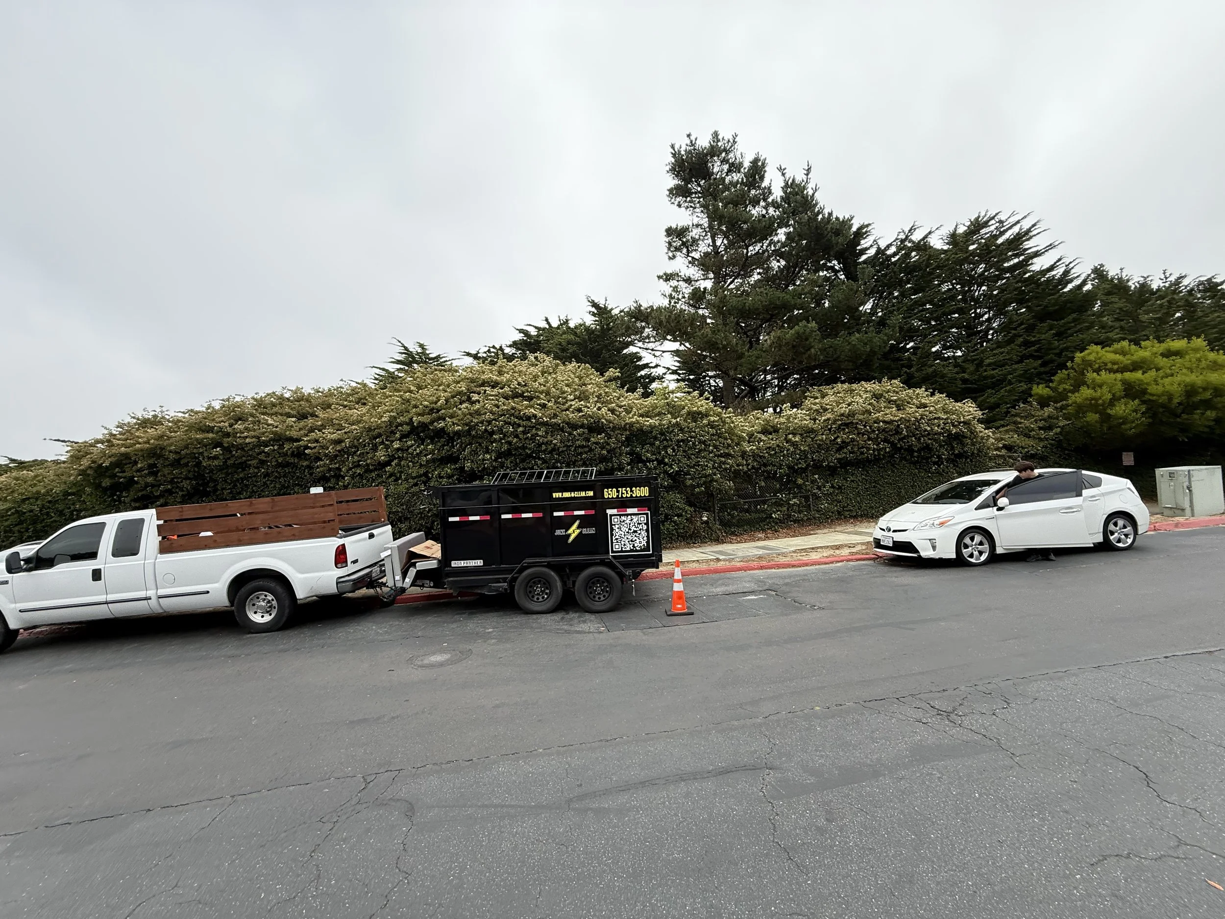 Parking lot with vehicles including a white pickup truck, a black trailer, and a white sedan, with trees and overcast sky in the background.