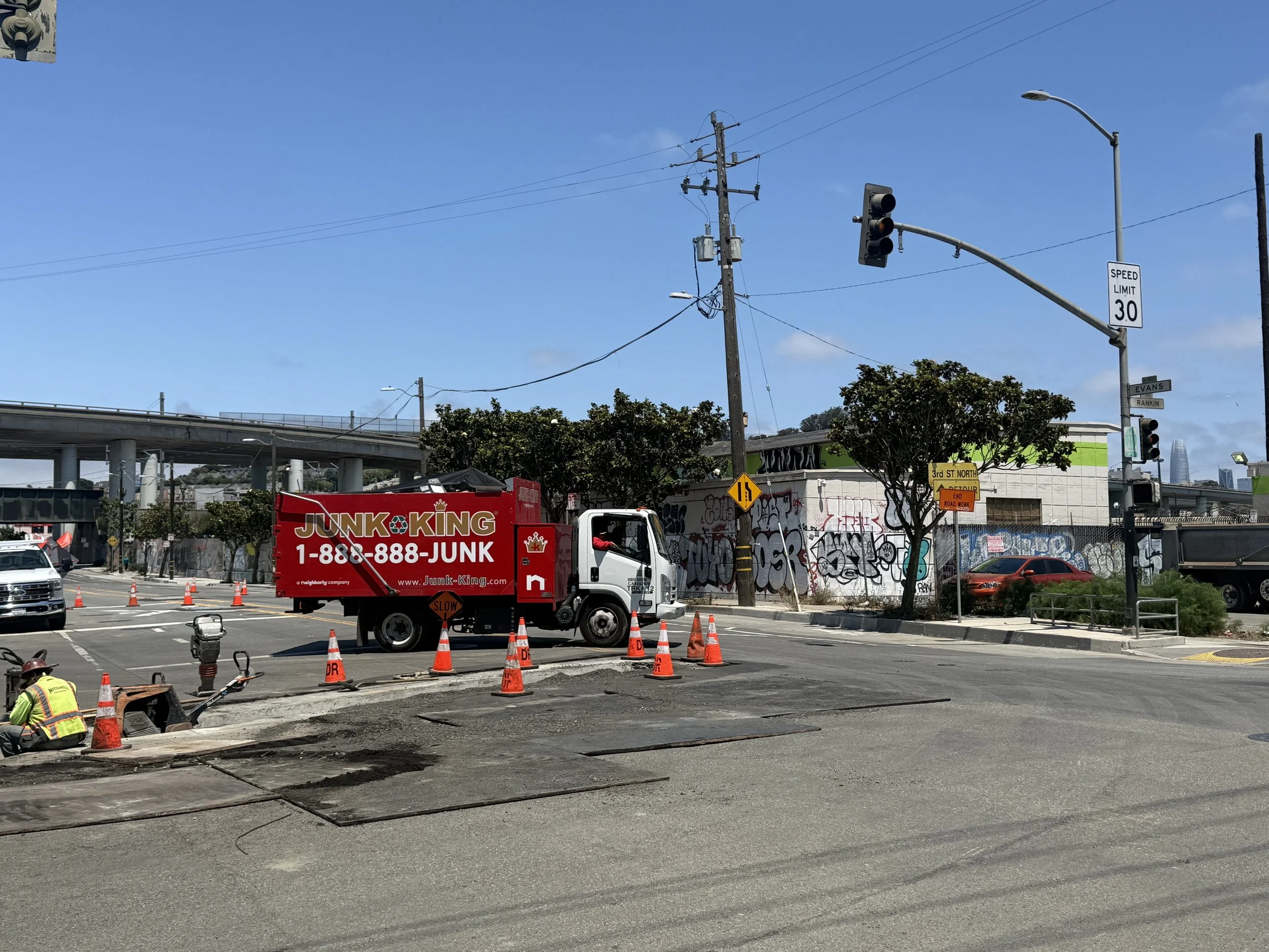 Construction workers and equipment working on a road repair project with orange cones, a second truck, and a billboard for Junk King, along with traffic lights, street signs, utility poles, and trees against a clear blue sky.