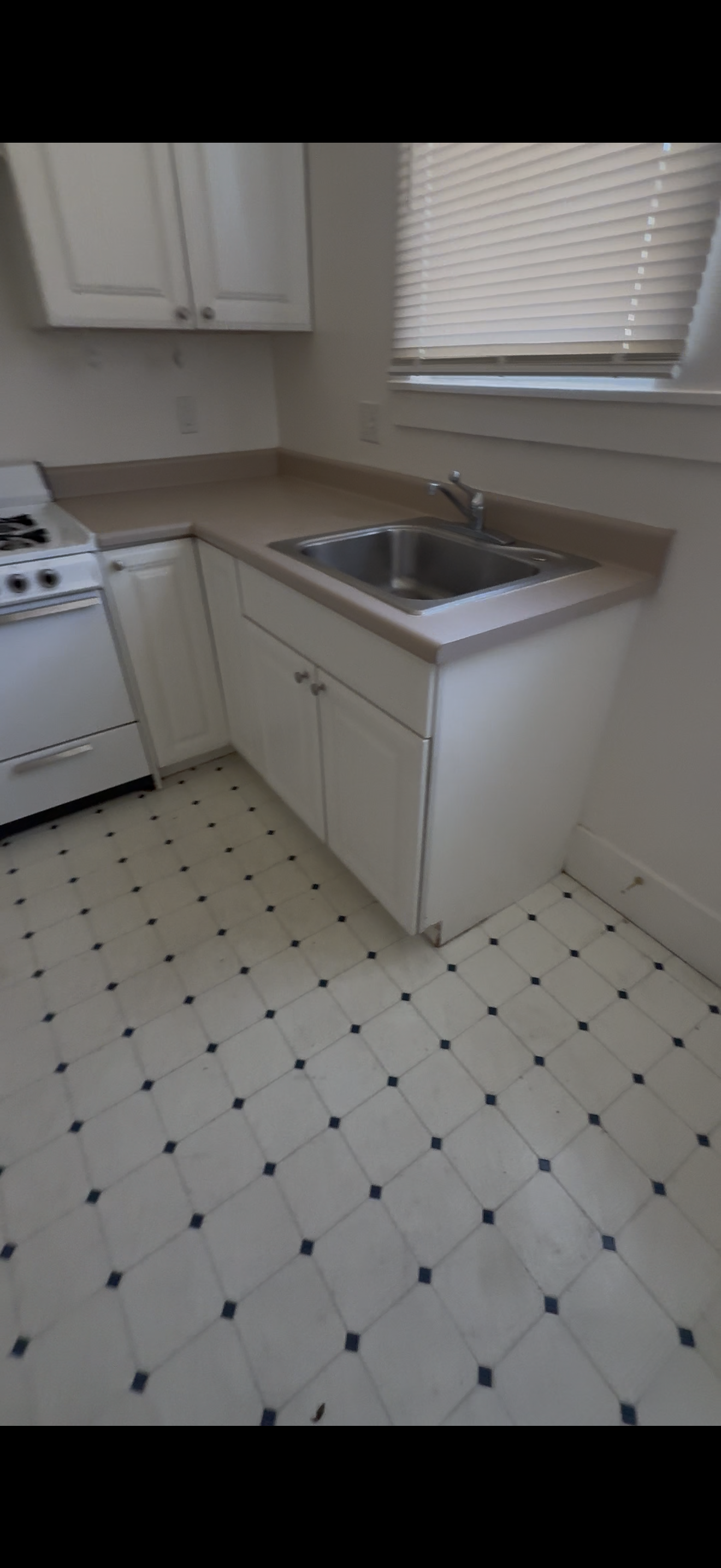 A small kitchen with white cabinets, a beige countertop, a stainless steel sink, a stove, a window with closed blinds, and a white floor with black diamond-shaped tiles.