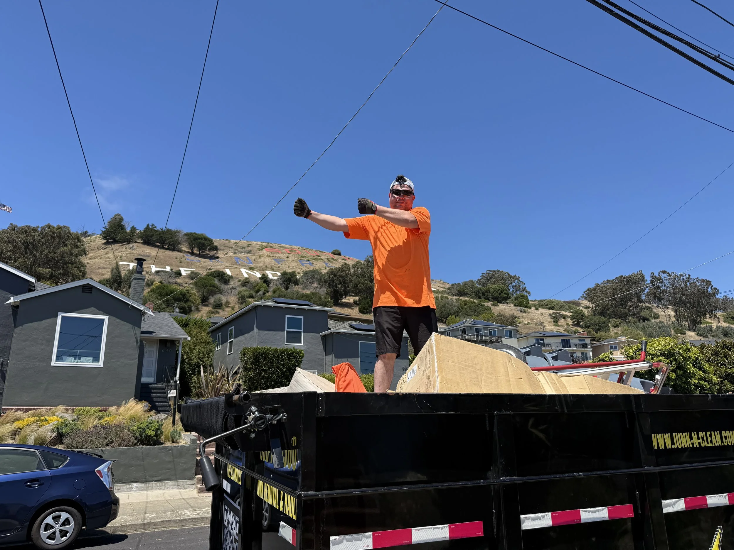 A man with sunglasses and a backwards cap, wearing an orange shirt, stands on a truck bed with a cloudy blue sky behind him. Houses and a hillside with trees are visible in the background.
