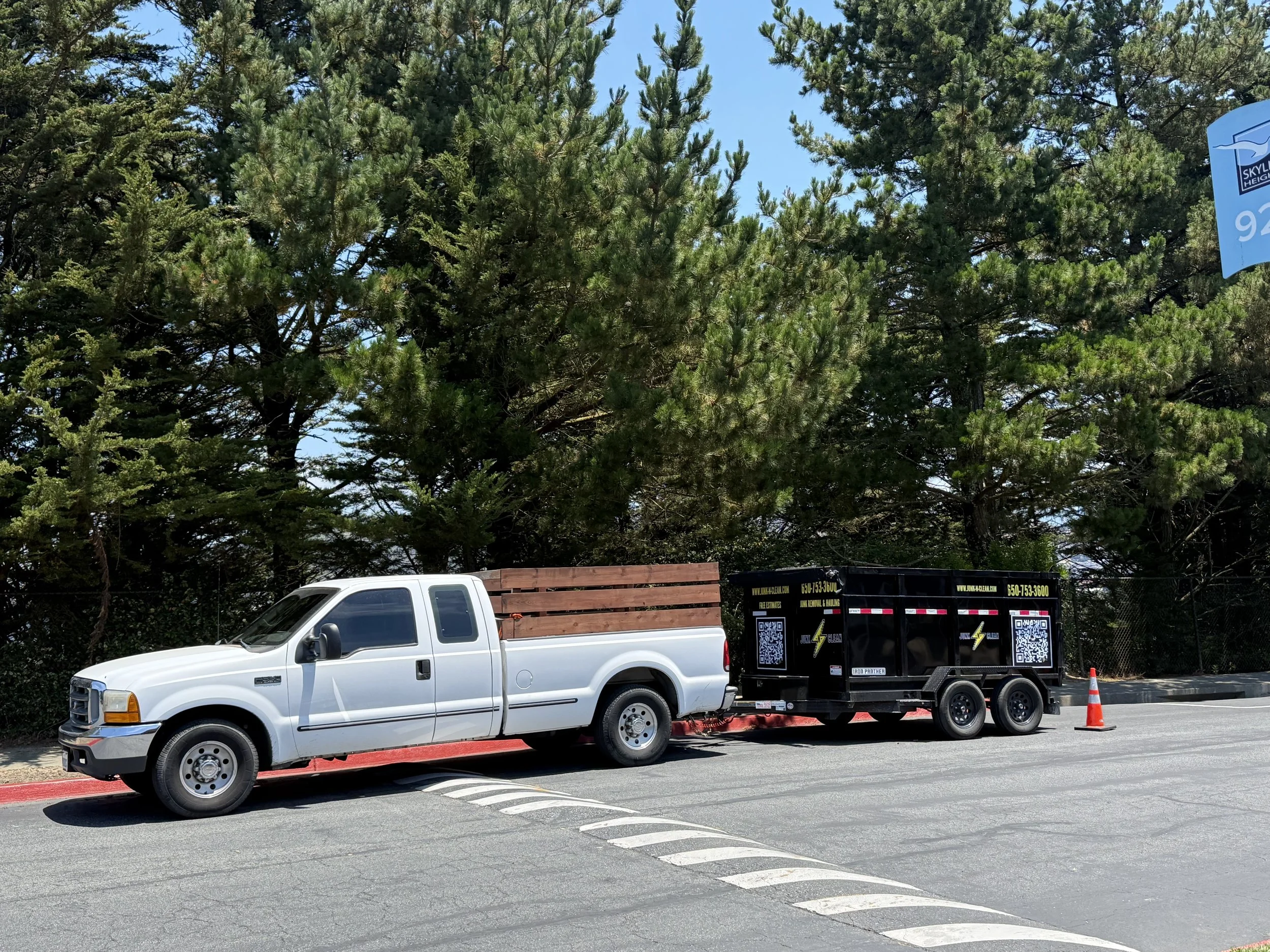 A white pickup truck with a black trailer attached, parked in a parking lot with a red curb. In the background, there are large green pine trees and a blue sky. There is a traffic cone near the truck.