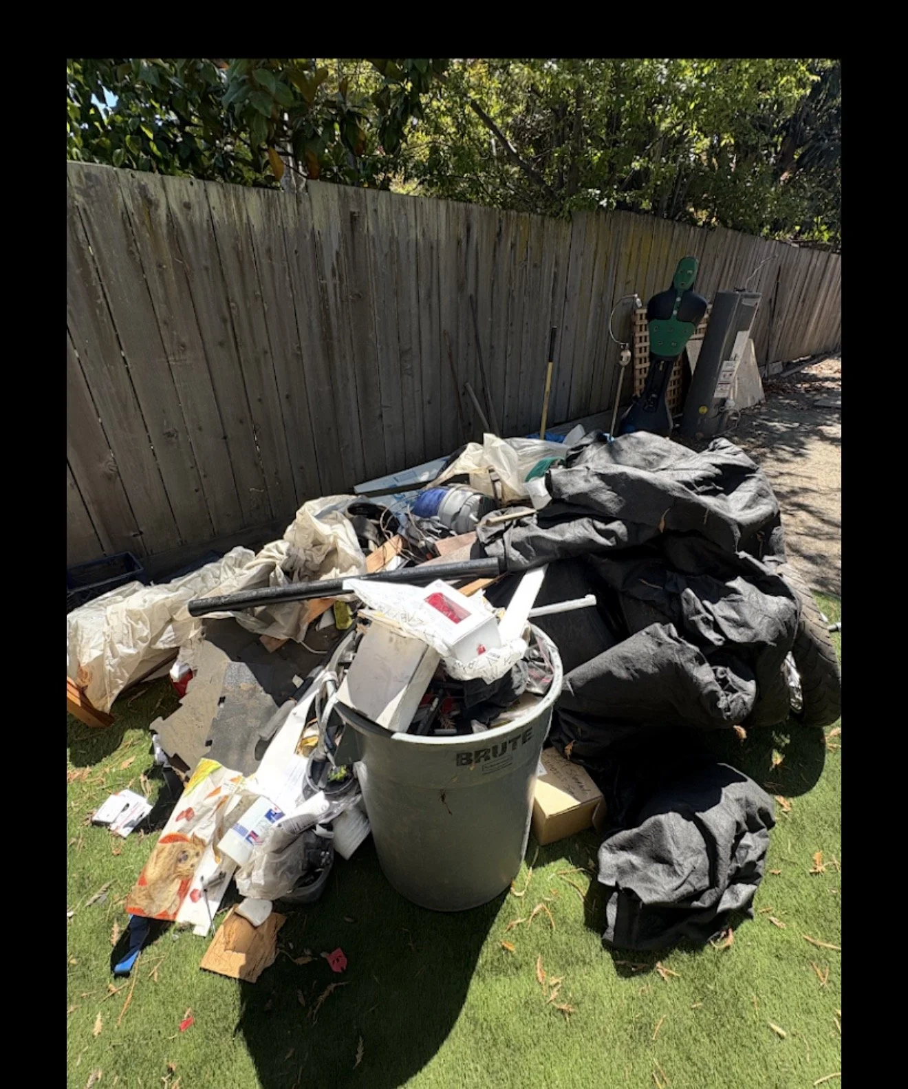 A pile of clutter and trash outdoors next to a wooden fence, including a large trash can, clothing, cardboard, and miscellaneous items, with trees and greenery in the background.