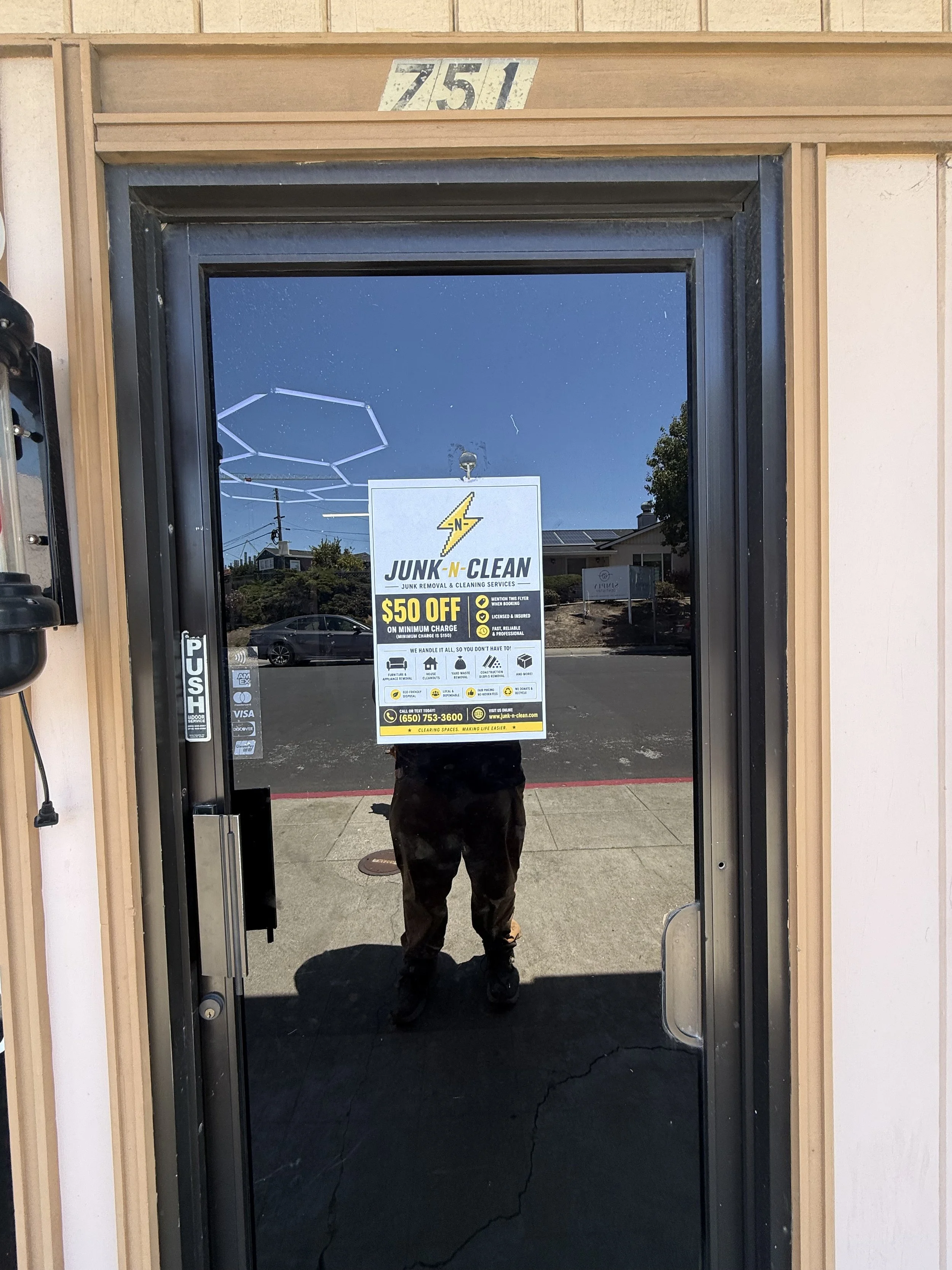 Reflected view of a person taking a photo inside a glass door with a sign advertising a junk removal business and a discount, and the outdoor scene including cars and trees visible in the reflection.
