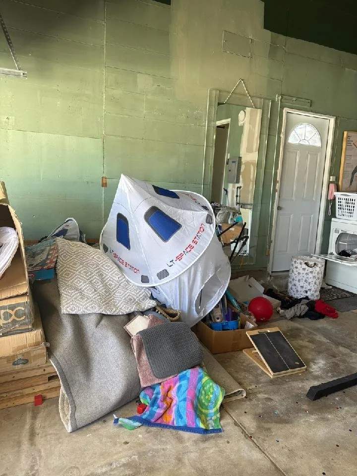 Cluttered garage or storage area with various items, including a Space Shuttle model, boxes, a laundry basket, and miscellaneous household objects.